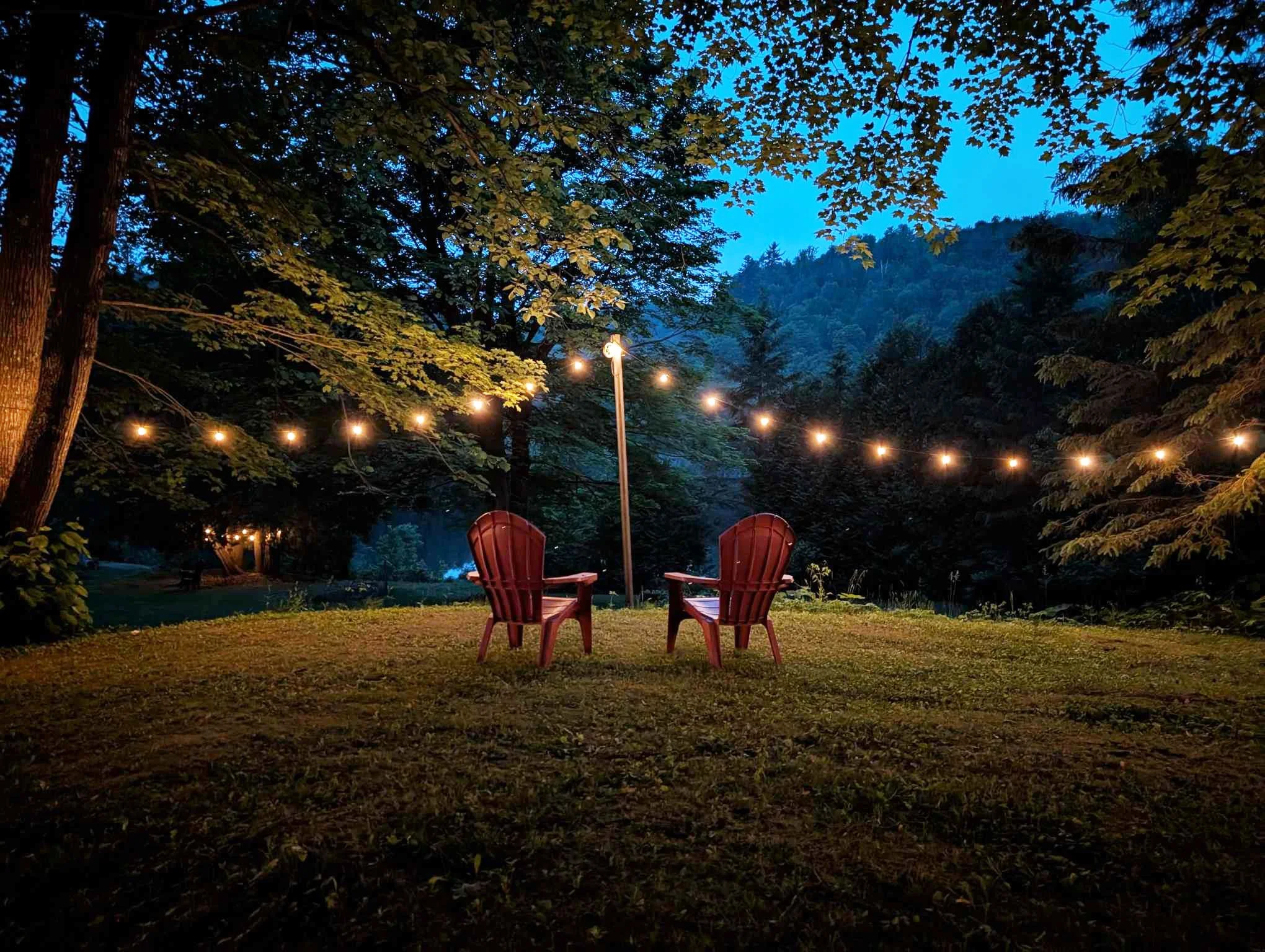 Two red Adirondack chairs facing each other on a grassy area at night, illuminated by string lights hung between trees in a wooded outdoor setting, with mountains in the background.
