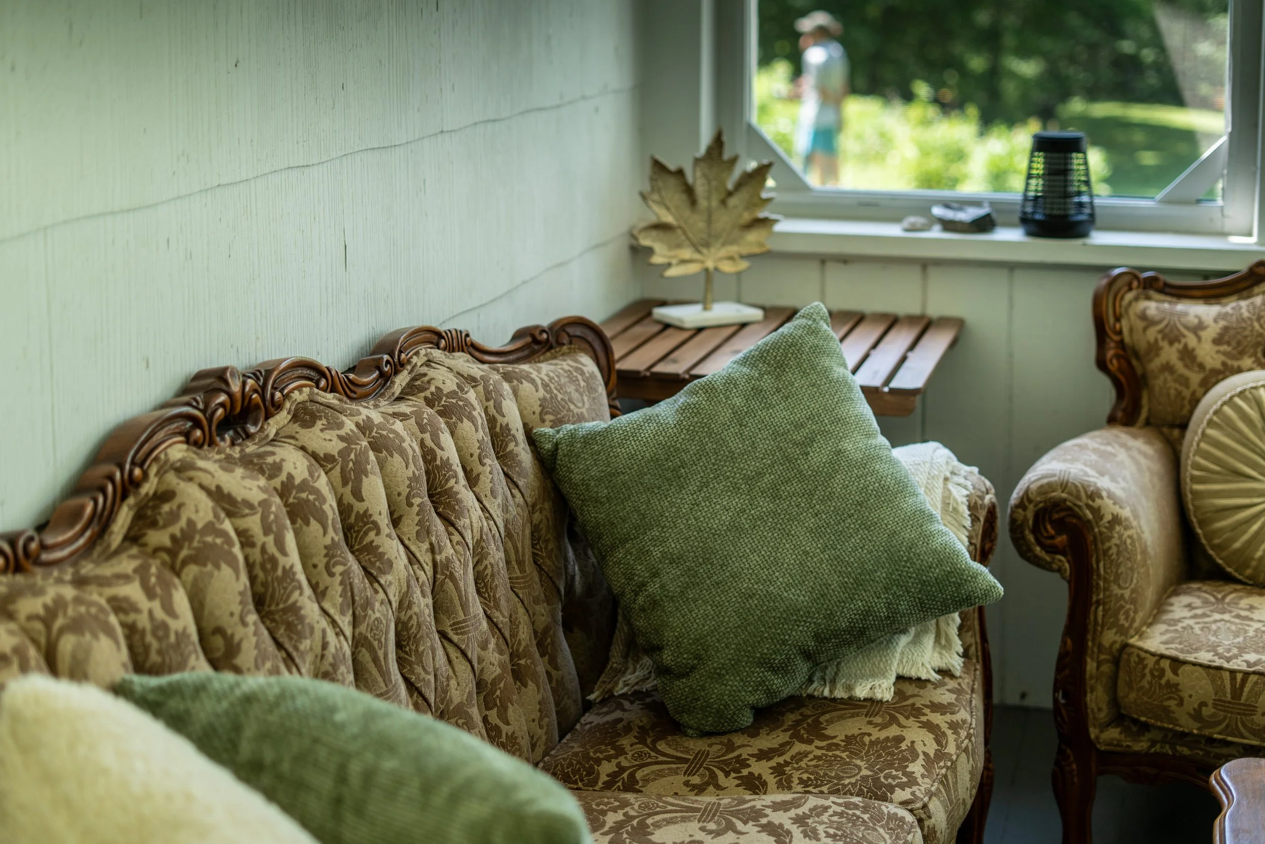 Vintage living room with floral patterned sofa and armchair, green pillows, small wooden side table, and window with greenery outside.