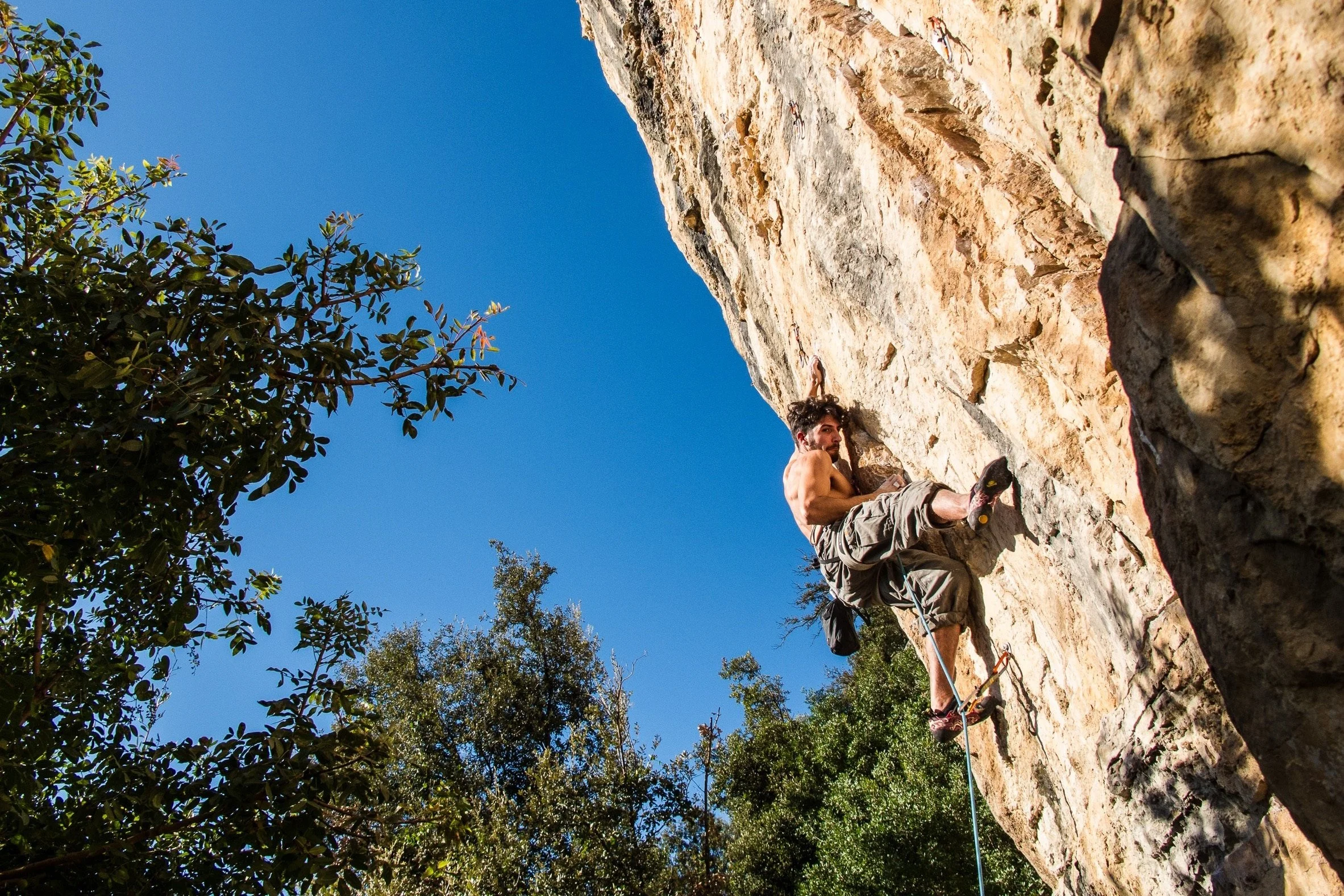 A man with a beard and curly hair shirtless, climbing a rocky outdoor cliff face with climbing shoes, harness, and rope, during daylight with clear blue sky and greenery around.
