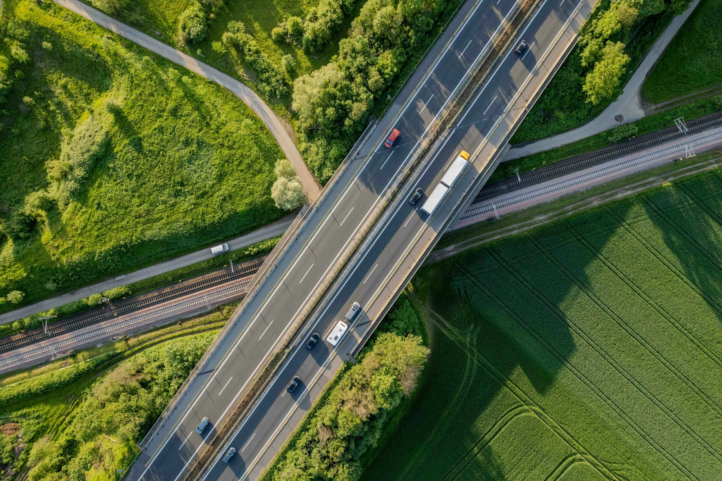 Una vista aérea de una autopista con varios autos y camiones, rodeada de áreas verdes con árboles y campos agrícolas.
