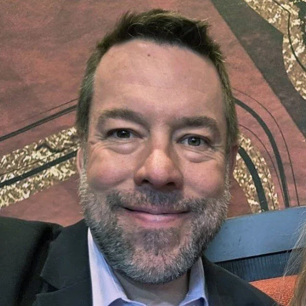 Close-up of Matthew DeRemer, with short brown hair and a beard, dressed in a suit, in an indoor setting with decorative flooring.
