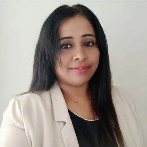 A woman with long black hair wearing a beige blazer and a black top, standing against a plain light-colored background.