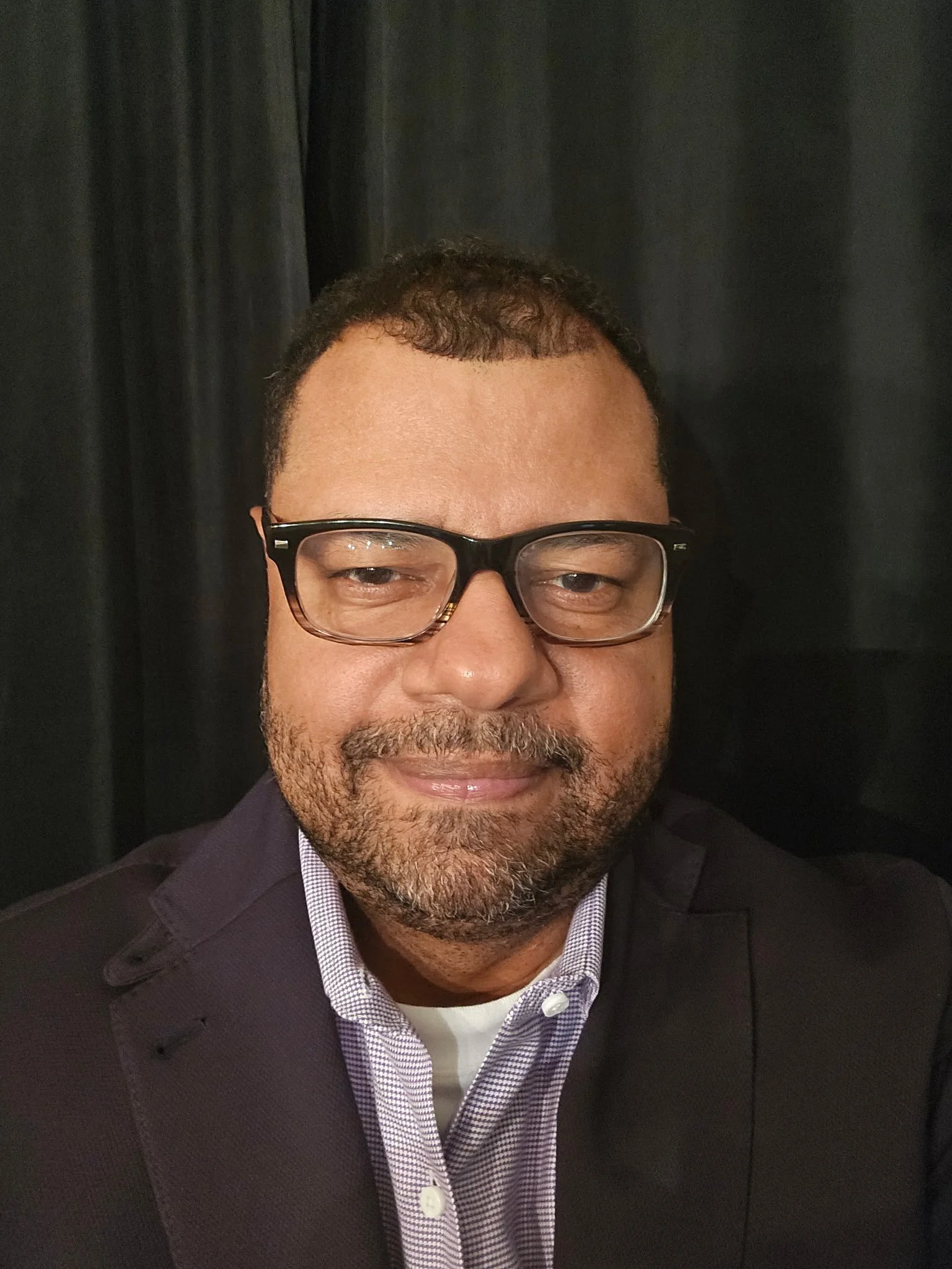 Dan Nunn with glasses, wearing a suit jacket and a collared shirt, smiling slightly against a dark background.