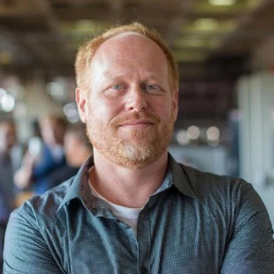 A man with a beard and short red hair smiling in a casual setting, possibly a cafe or restaurant.