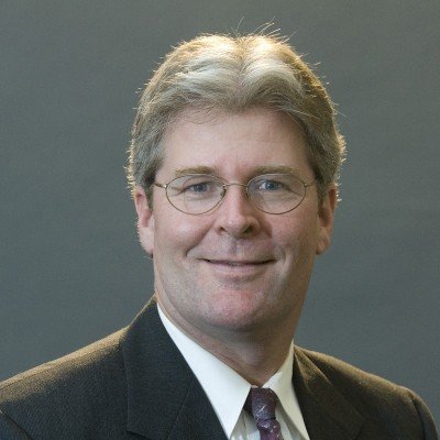 Bradley George, s, wearing a dark suit, white shirt, and patterned tie, smiling at the camera.
