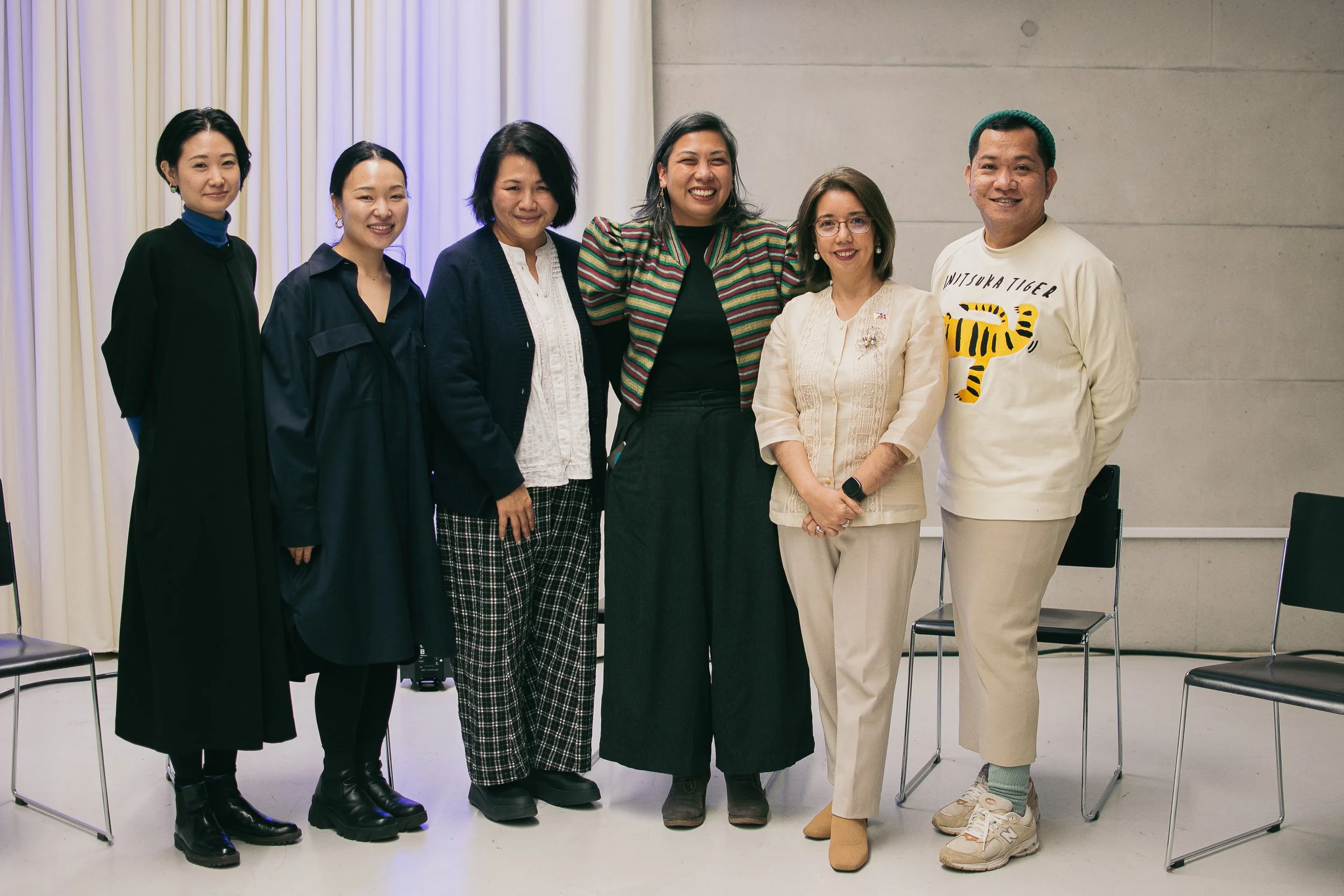 Six diverse women and one man standing together indoors in front of beige curtains and a gray wall, smiling.