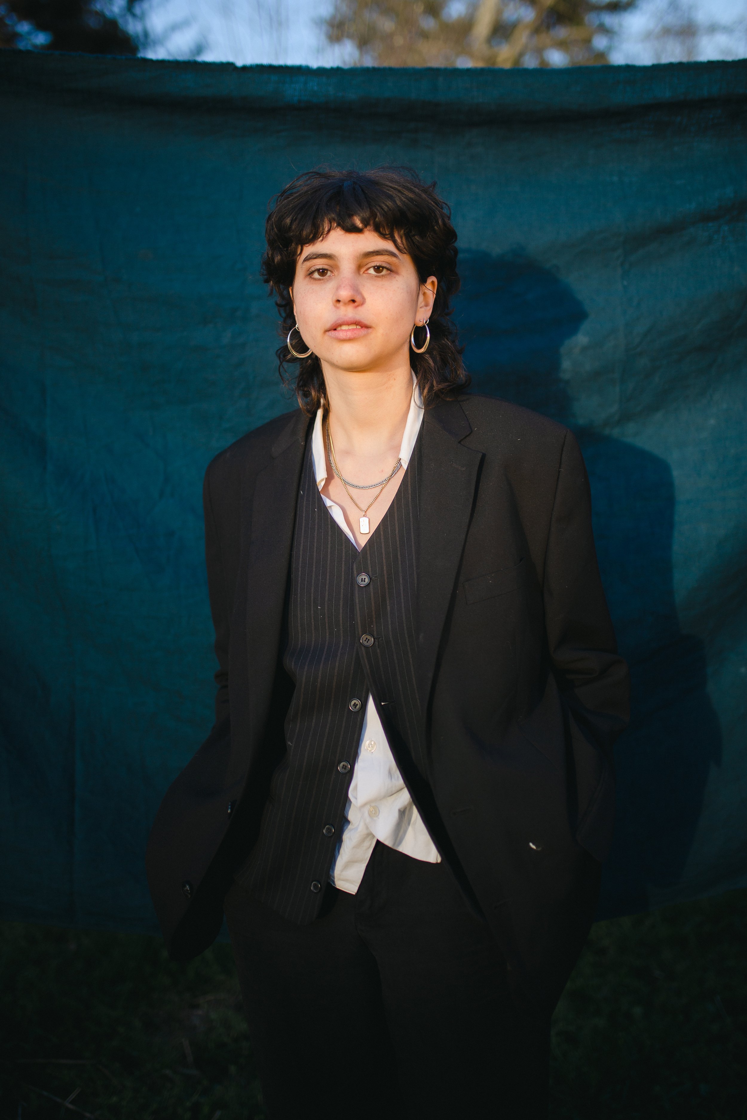 A woman with short dark curly hair wearing a black blazer, black pinstripe vest, white shirt, multiple necklaces, and hoop earrings standing outdoors in front of a blue tarp.