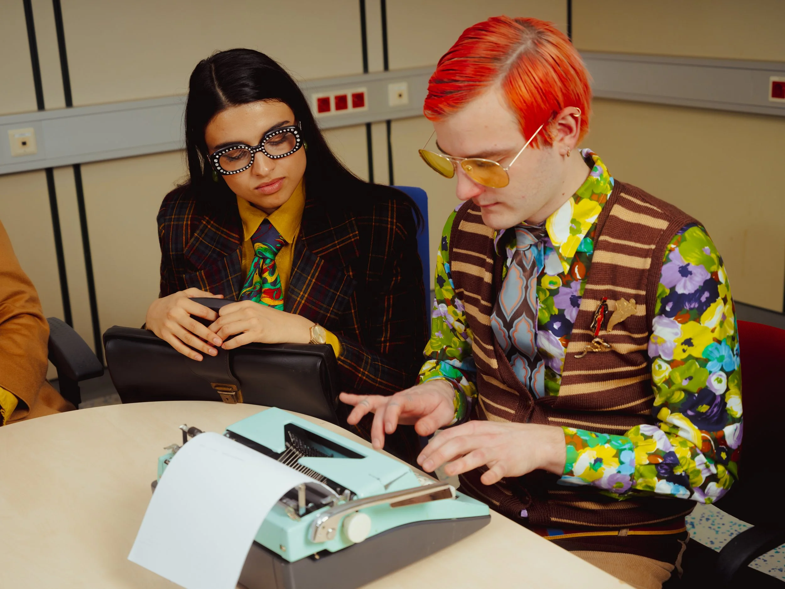 Two people dressed in colorful, retro-style clothing sitting at a table with a vintage typewriter and a sheet of paper. The woman has long dark hair, glasses, and a plaid blazer, while the man has bright orange hair, yellow-tinted sunglasses, and a f