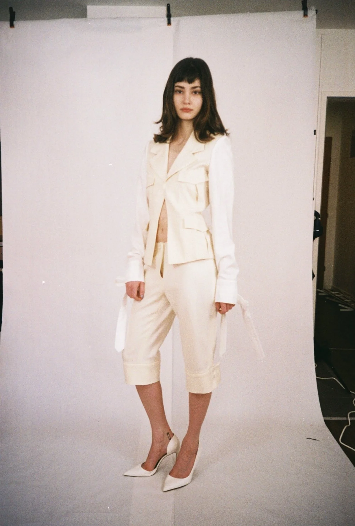 A woman with shoulder-length brown hair and bangs wearing a cream-colored suit with cropped pants and high heels, standing in front of a plain white backdrop.