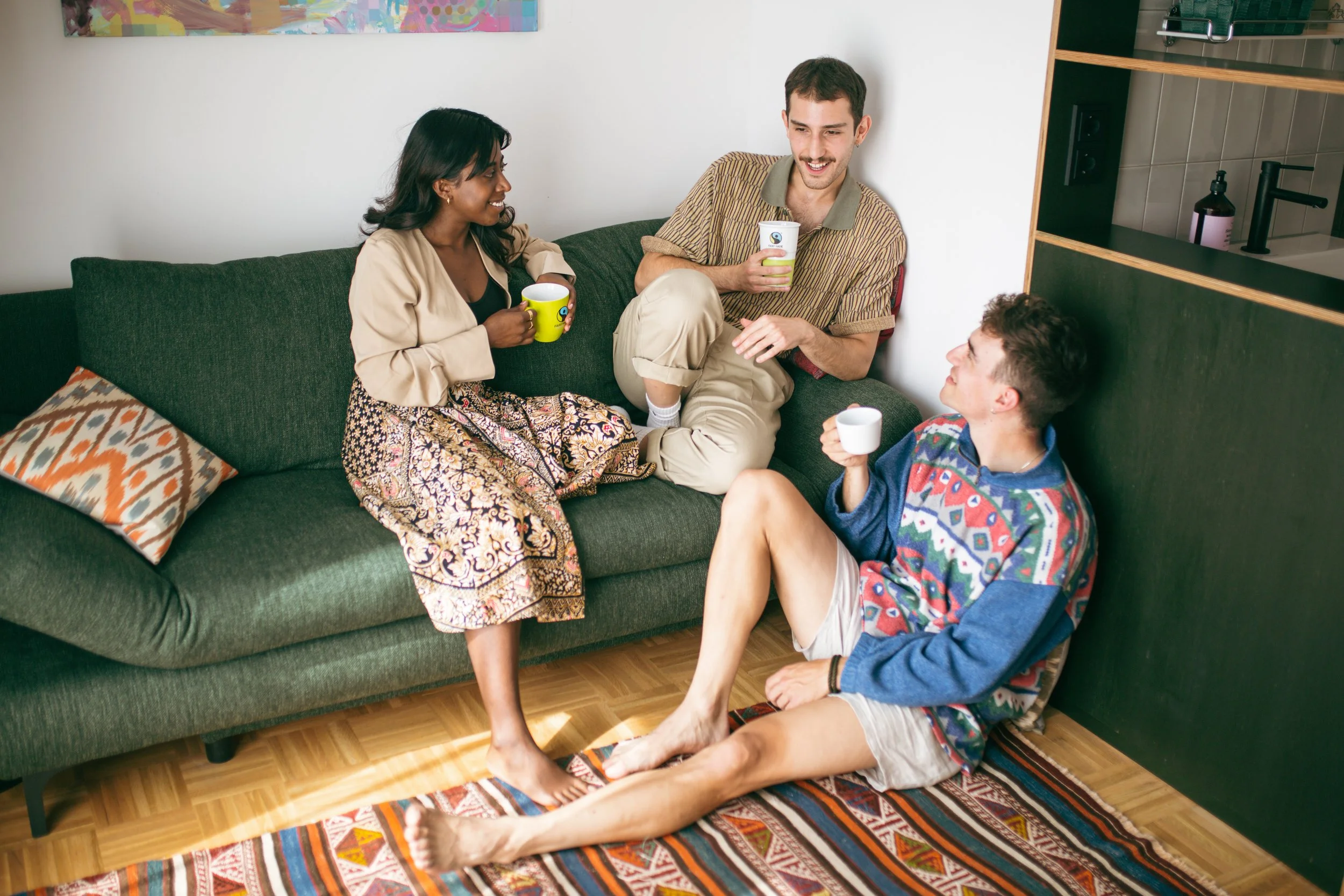 Four young adults are having a conversation in a cozy living room. Two of them are sitting on a dark green couch, while one person is sitting on the floor with legs outstretched. They are all holding cups and smiling at each other.