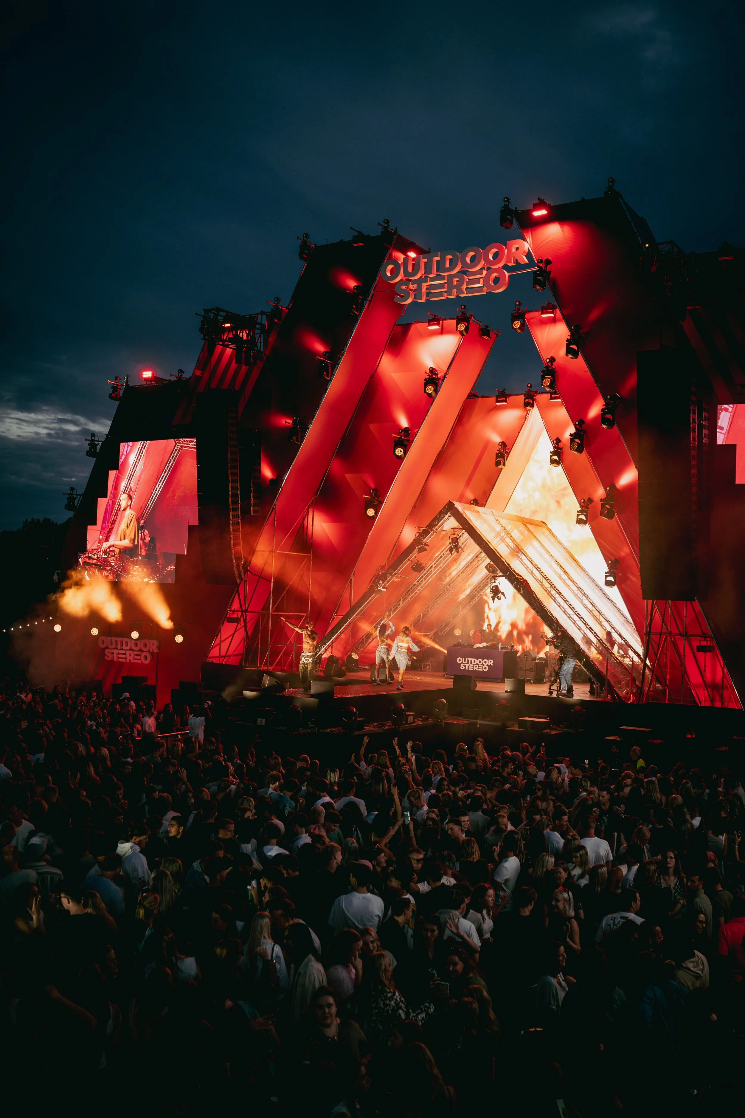 A large outdoor concert stage illuminated with red lights, featuring the words 'OUTDOOR STEREO' at the top. A DJ is performing on the stage surrounded by a seated audience during the evening.