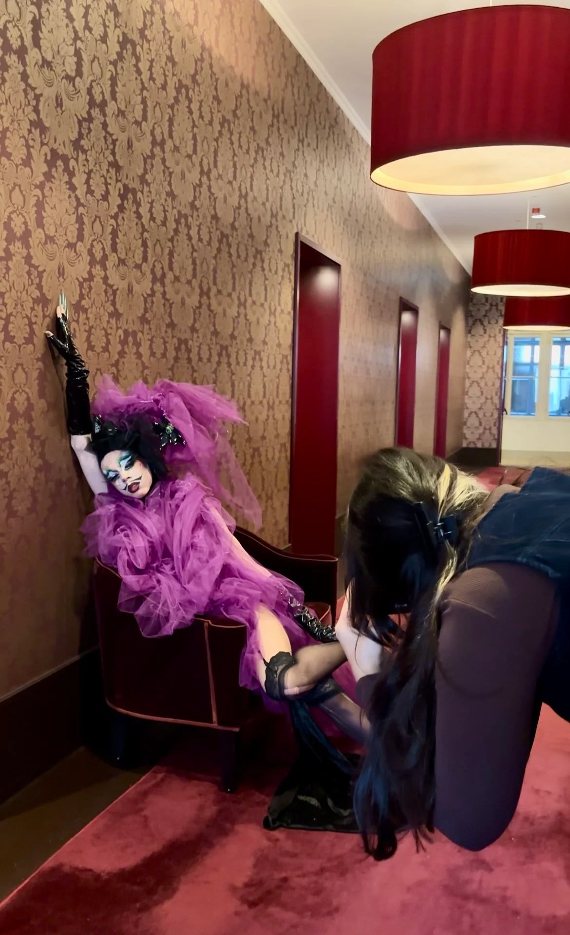 A person dressed in elaborate purple and black gothic clothing, with dramatic makeup, is seated on a purple chair against a patterned wall. Another person with long hair, wearing headphones, is adjusting their boots.