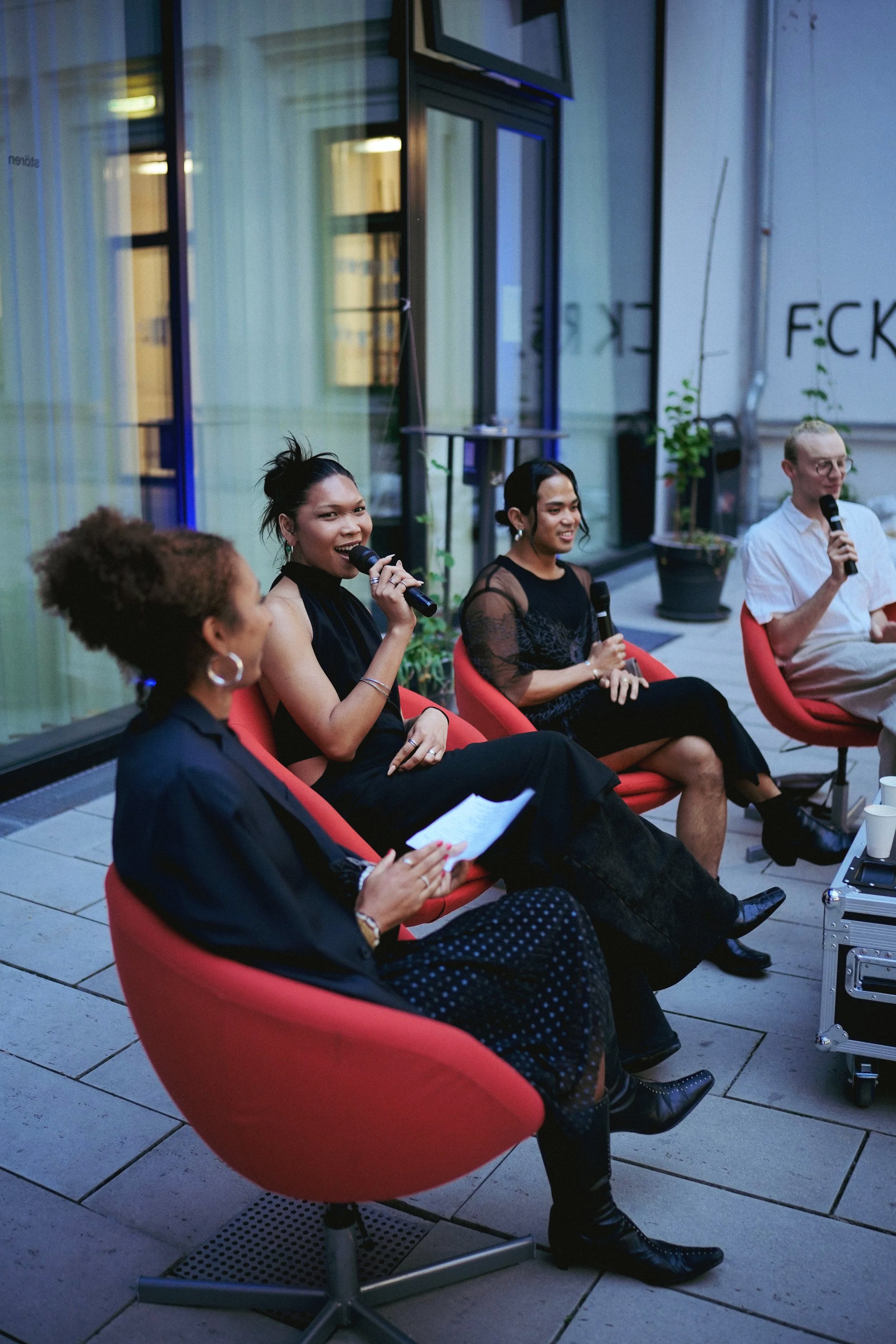 Four people sitting on red chairs in a panel discussion, with three women and one man, holding microphones, in a modern indoor setting with large windows and potted plants.