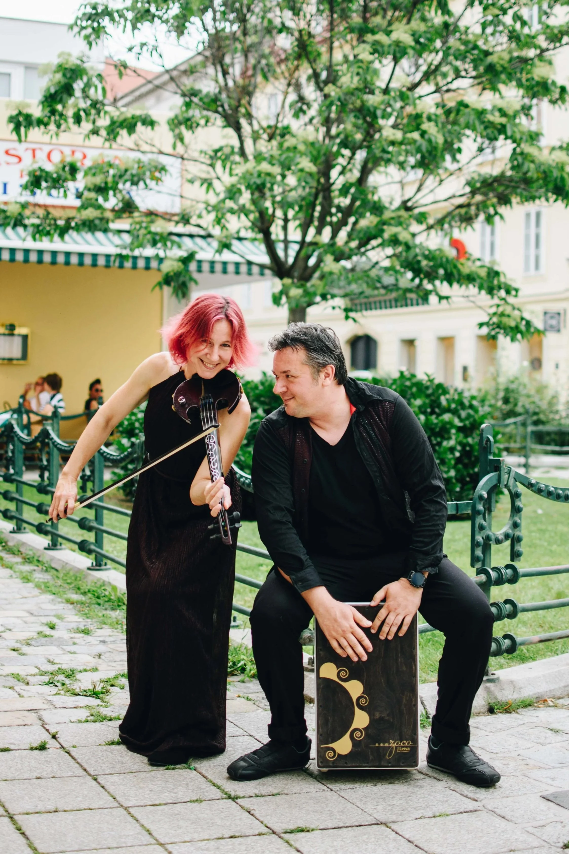 A woman with pink hair playing a violin and a man sitting on a cajón drum are performing outdoors on a cobblestone path, with a tree and buildings in the background.