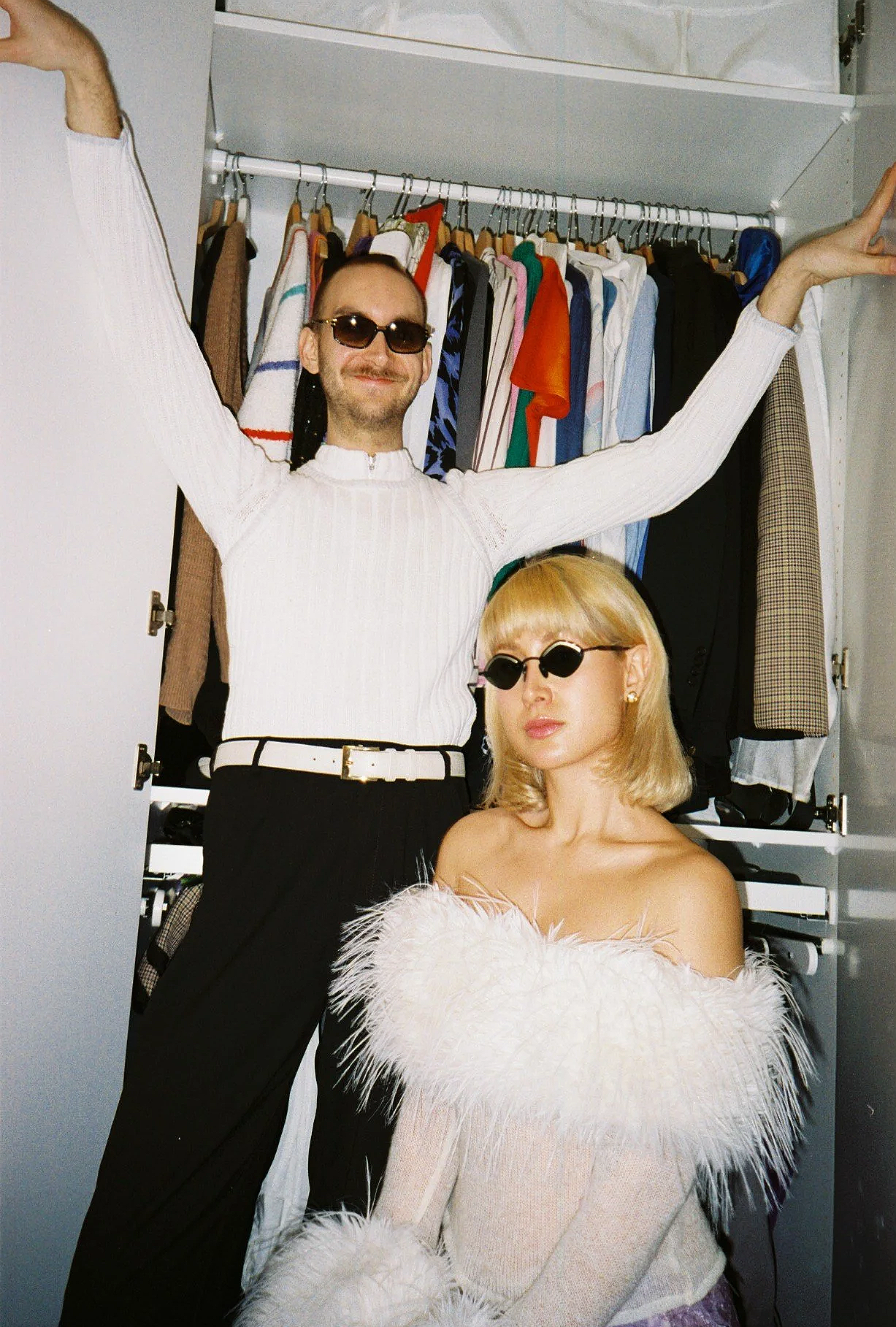 A man and woman posing inside a closet filled with hanging clothes. Both are wearing small, dark sunglasses. The man has a beard and long hair, and the woman has blonde hair with bangs and is wearing a fuzzy off-shoulder top.