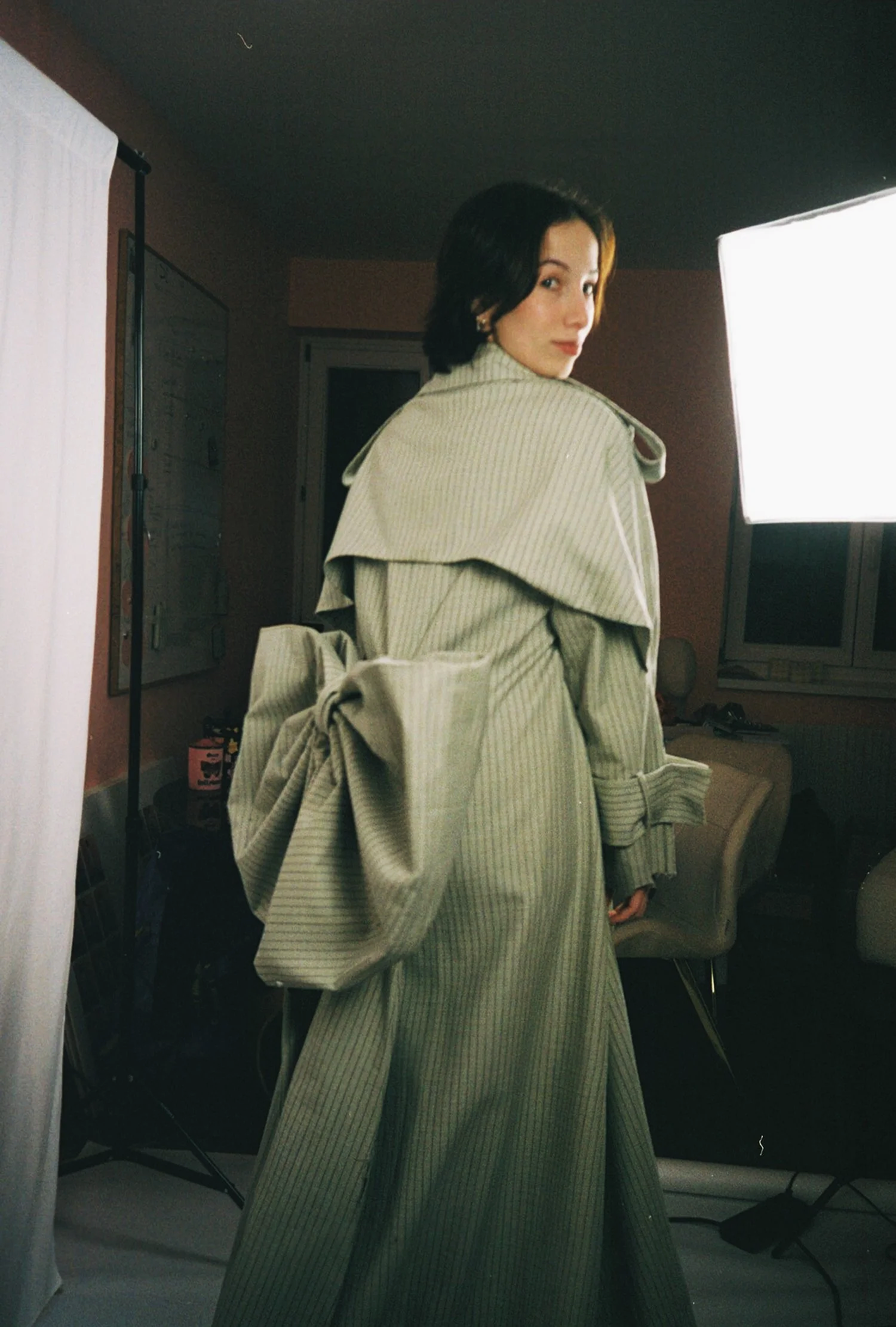 A woman in a long, beige striped trench coat, standing in a room with professional lighting equipment, looking over her shoulder.