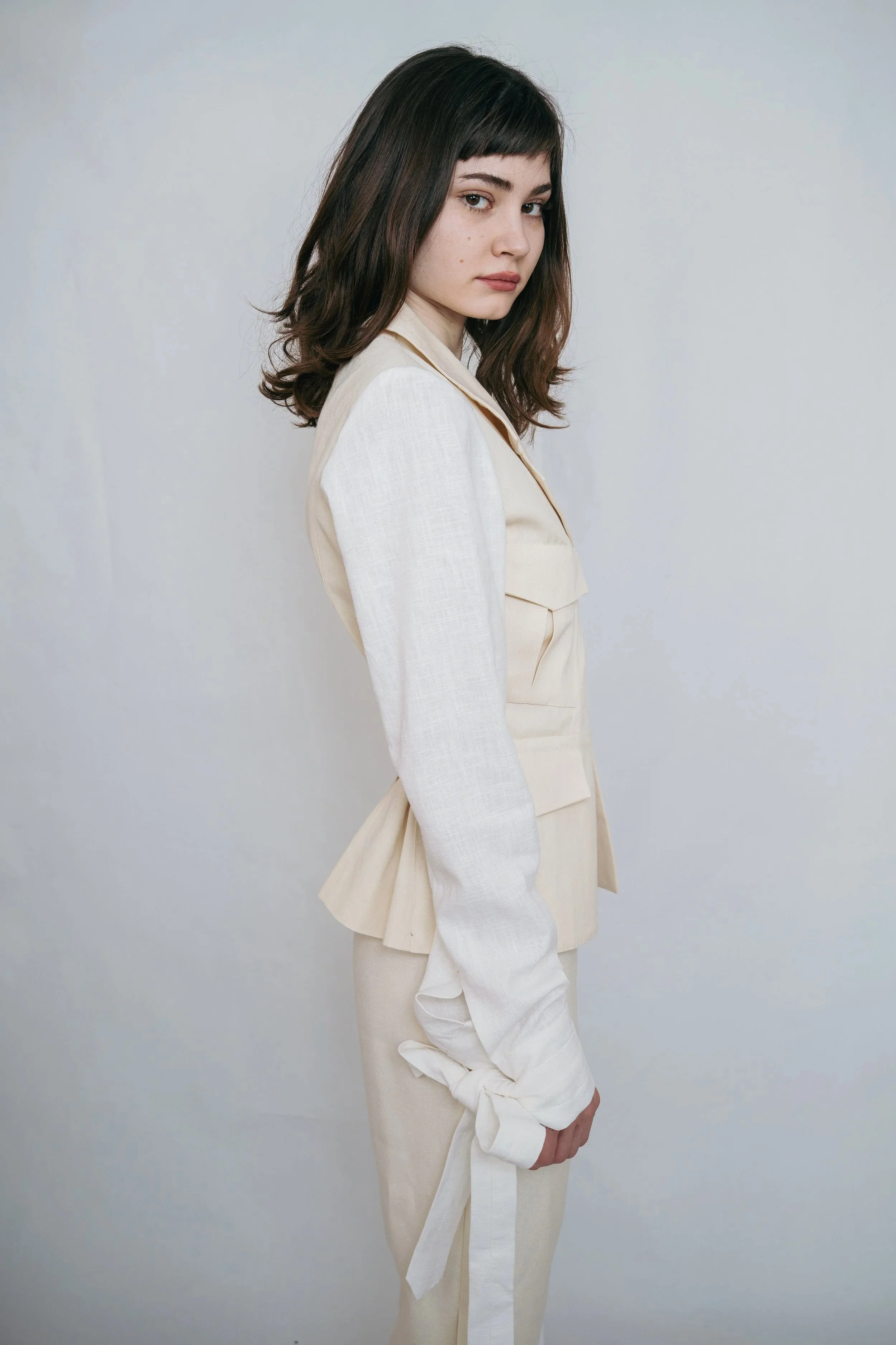 A young woman with dark brown hair, wearing a cream-colored blazer and matching pants, standing against a plain light-colored background.