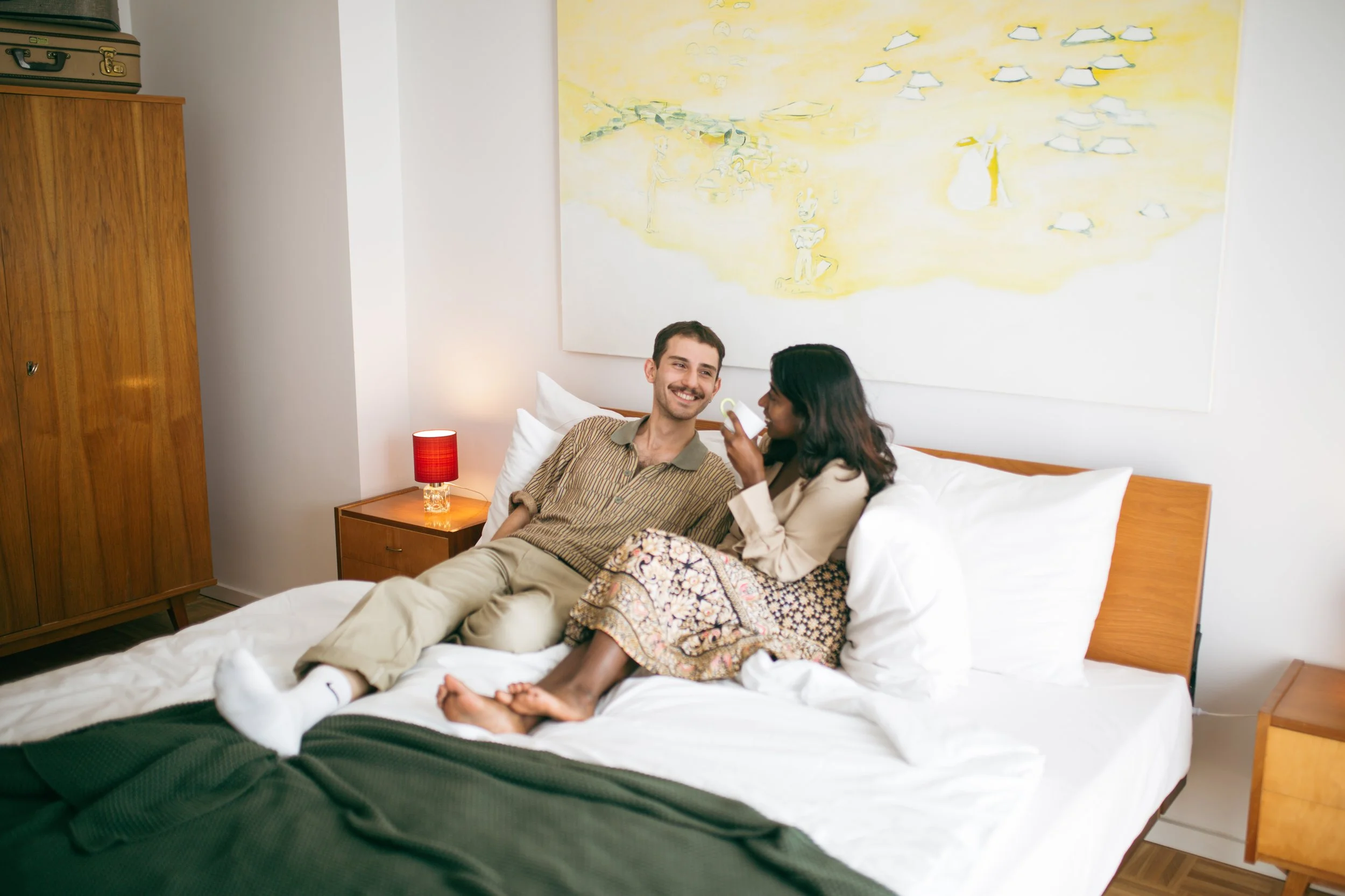 A man and woman sitting on a bed in a bedroom, smiling at each other, woman applying lotion on man's face, with furniture and a painting on the wall.