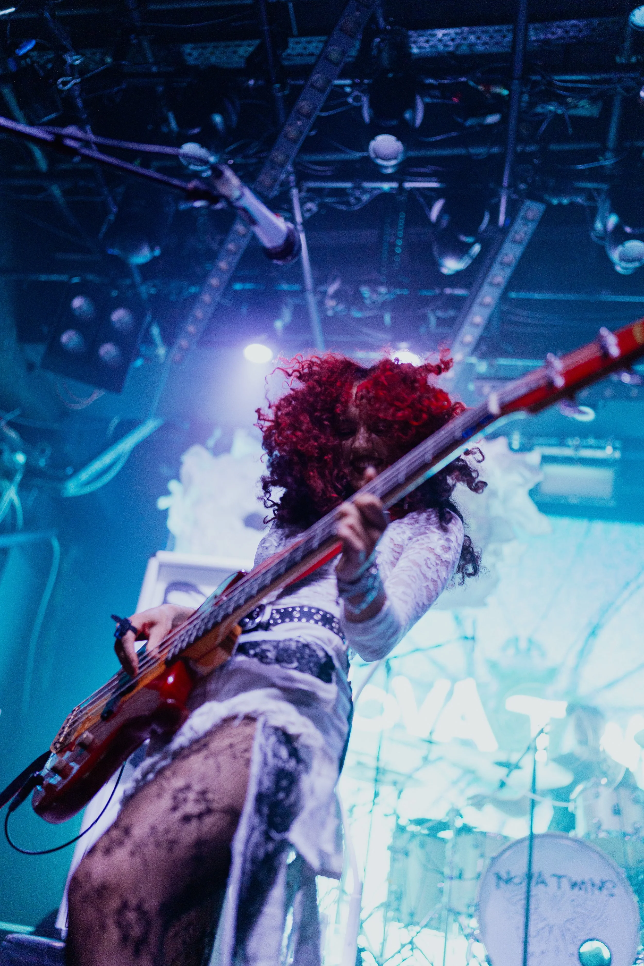 Female musician with curly red hair playing bass guitar on stage with bright blue and white lights behind her.