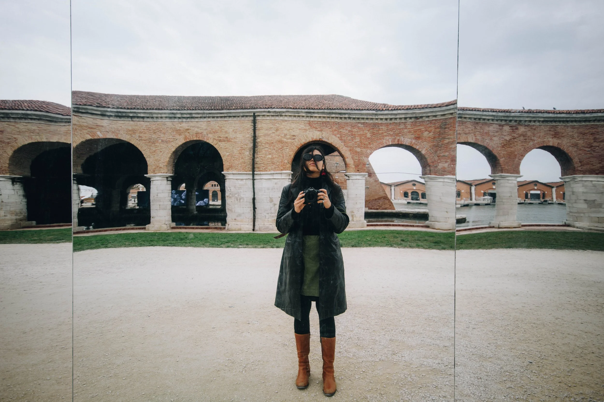 A woman taking a selfie in front of a large, ornate mirror with multiple panels, reflecting an old brick building with arches and a cloudy sky.