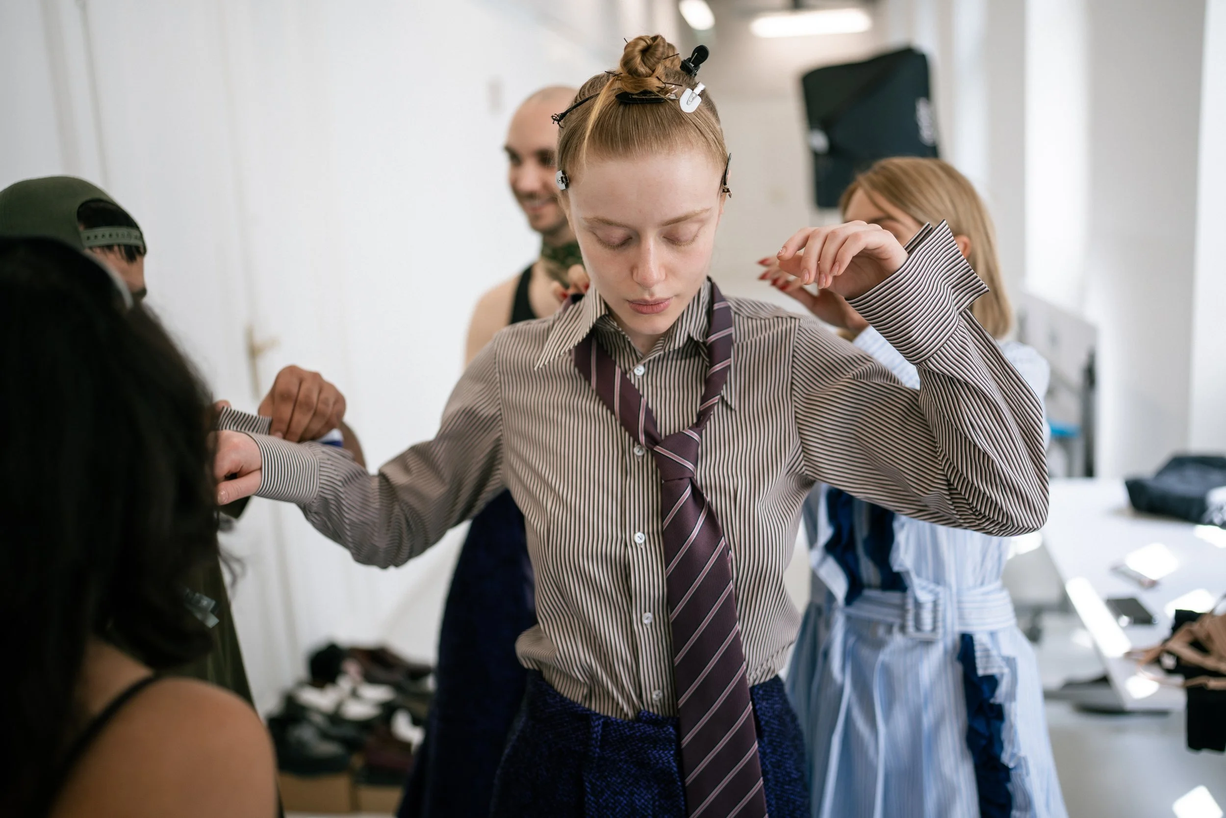 Young woman with light skin and blonde hair in a top knot adjusting her striped shirt and maroon tie backstage at a fashion show with other women and designer shoes in the background.