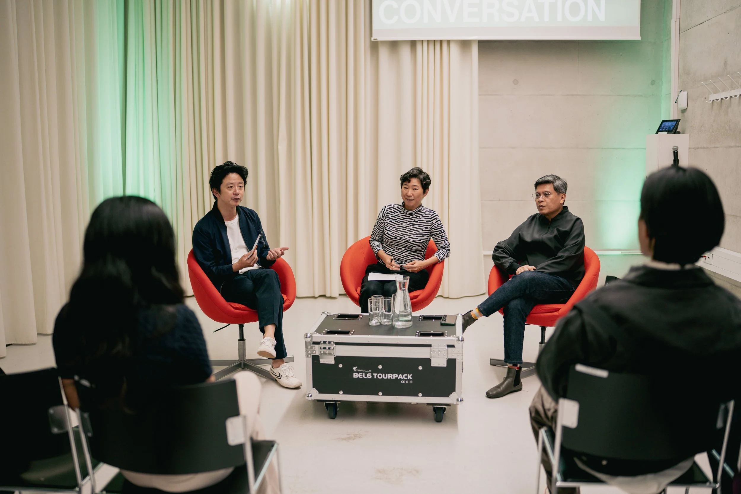 A panel discussion with three speakers seated in red chairs, engaging with an audience in a modern, well-lit room. A laptop, glasses of water, and a pitcher are on a black equipment box in front of them. A large screen behind displays the word 'CONVE