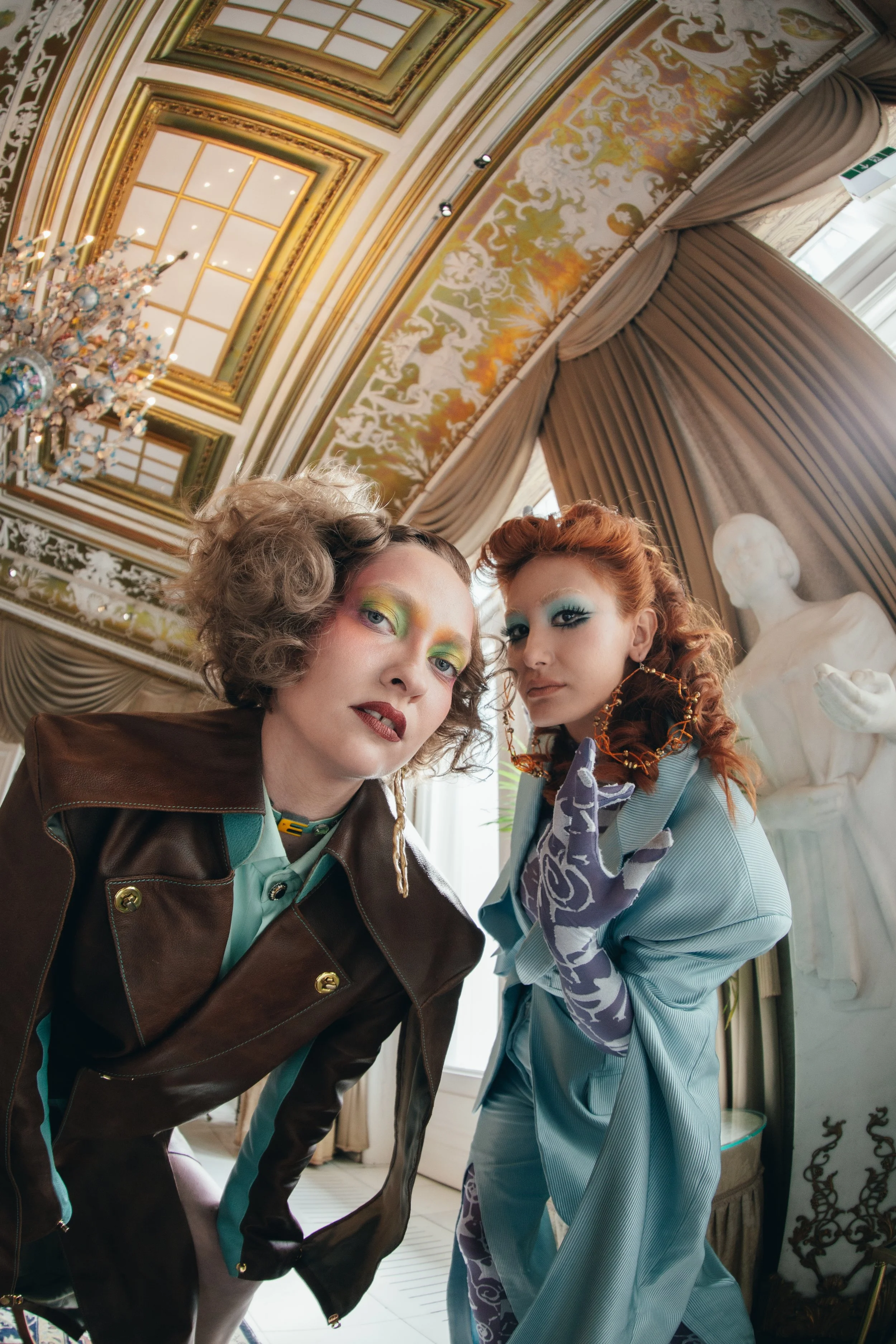 Two women with colorful makeup and fashion-forward clothing pose closely in an ornate room with a chandelier and classical sculpture.