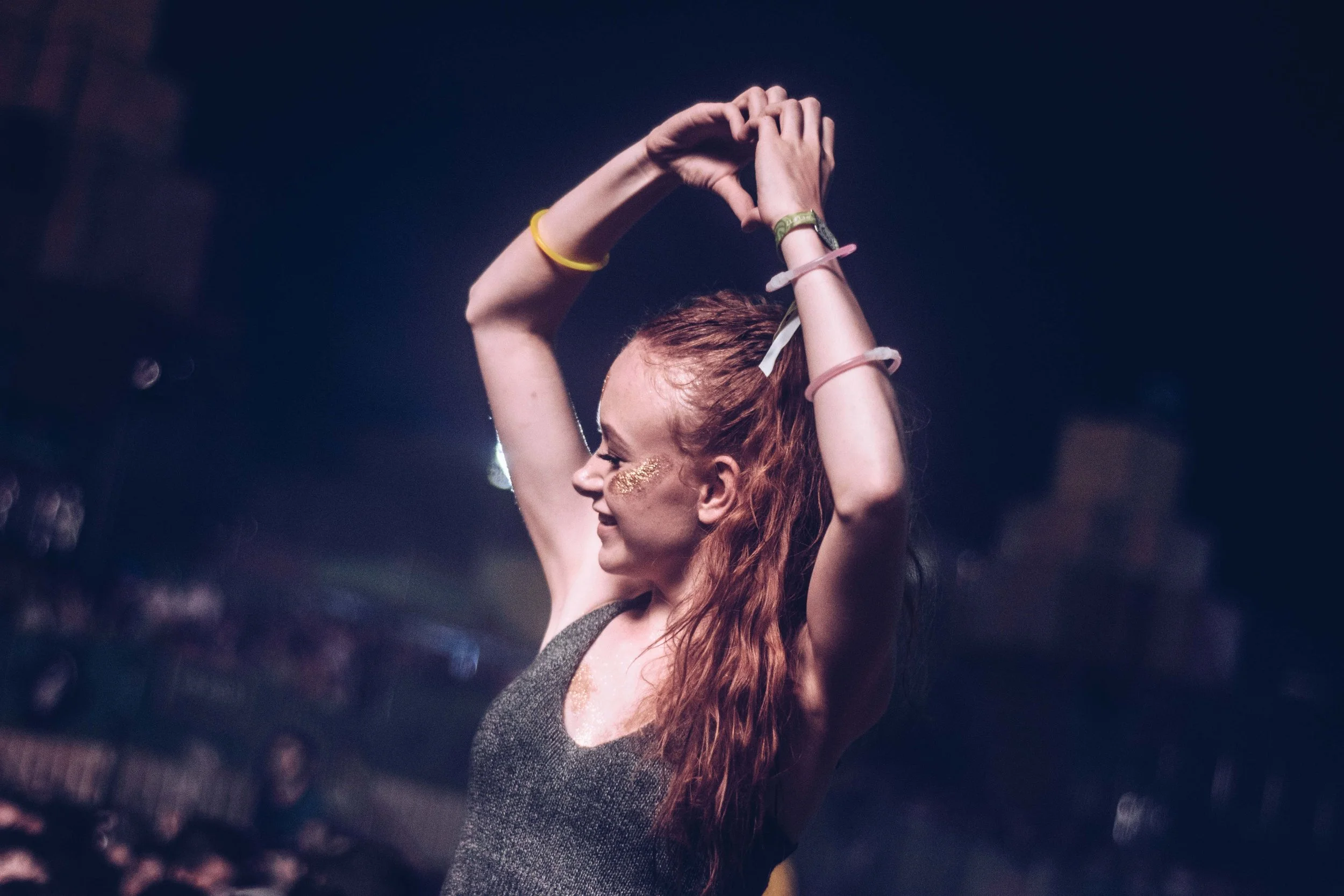 A woman with red hair dancing with her arms raised above her head in a dark club or concert setting, wearing a sleeveless top and bracelets, with glitter on her face.