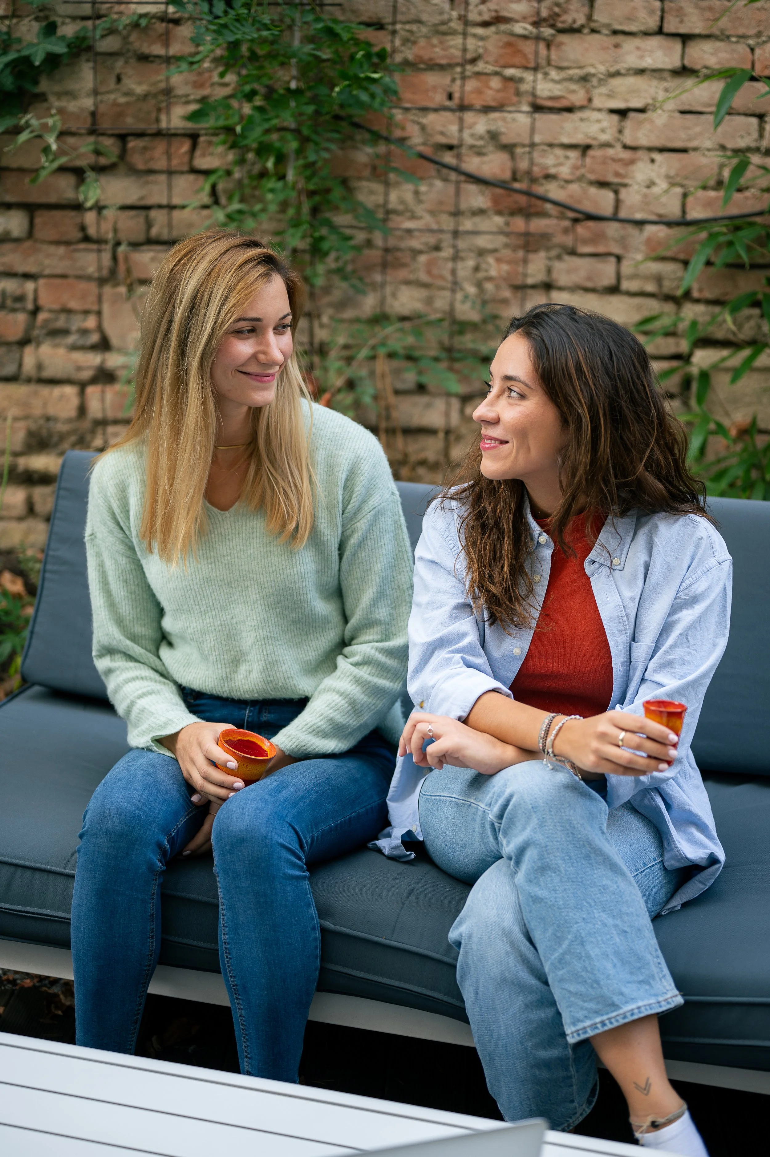 Two women sitting on a gray outdoor sofa, smiling at each other, one holding a small bowl, with a brick wall and green plants in the background.