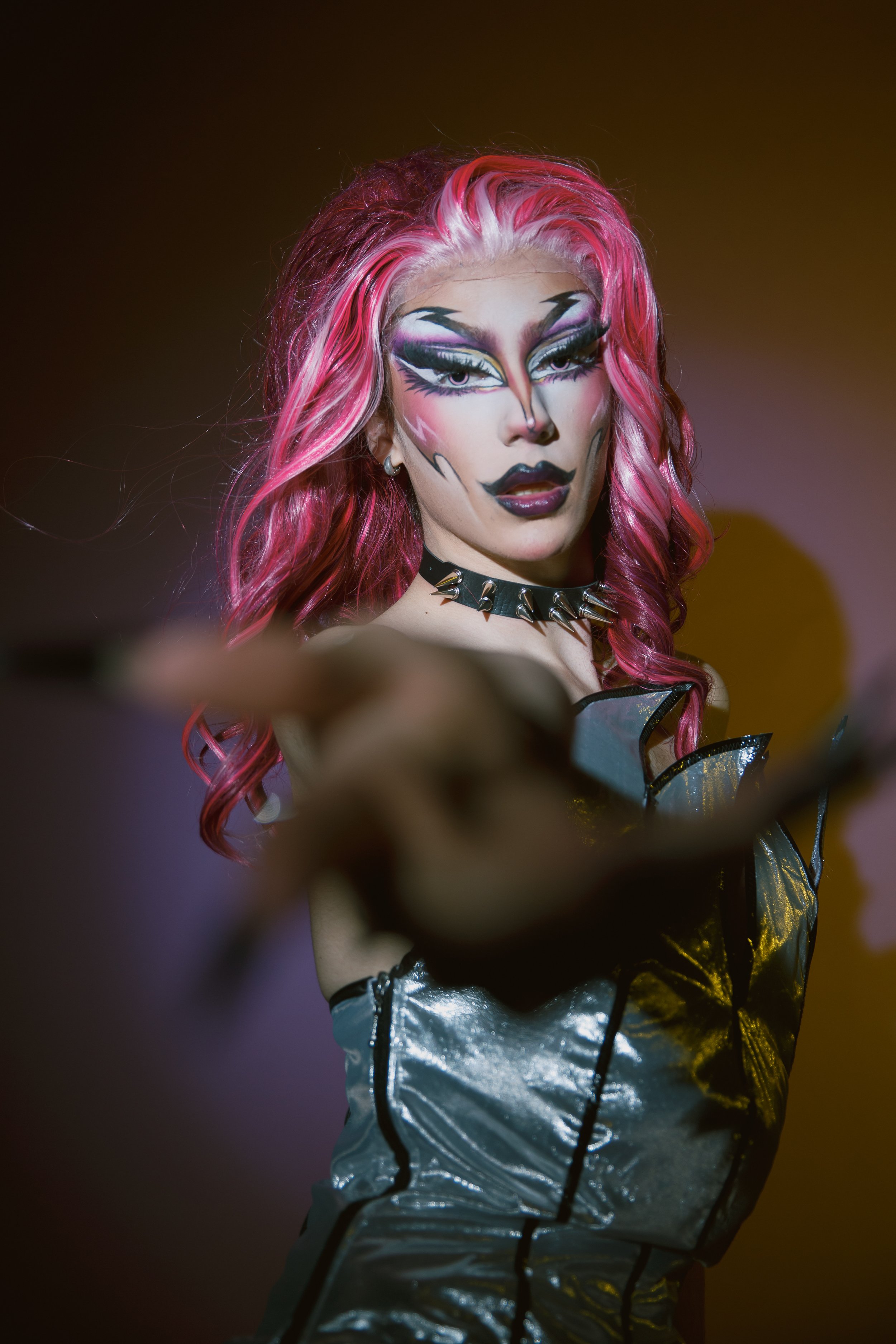 A drag performer with pink and purple hair, dramatic makeup, and a fierce expression, pointing forward.