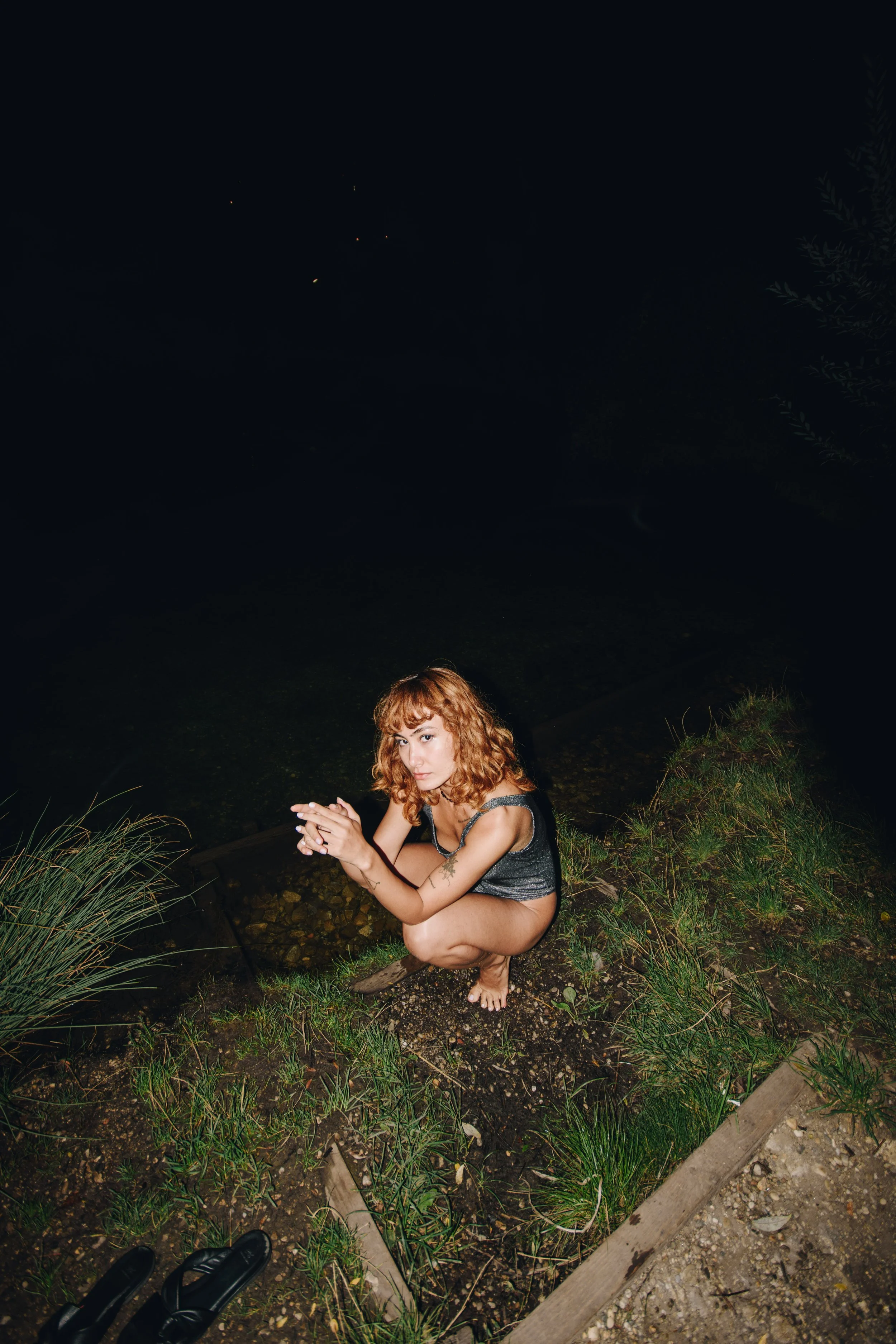 A woman with curly red hair crouching on a grassy patch near water at night, looking at the camera.