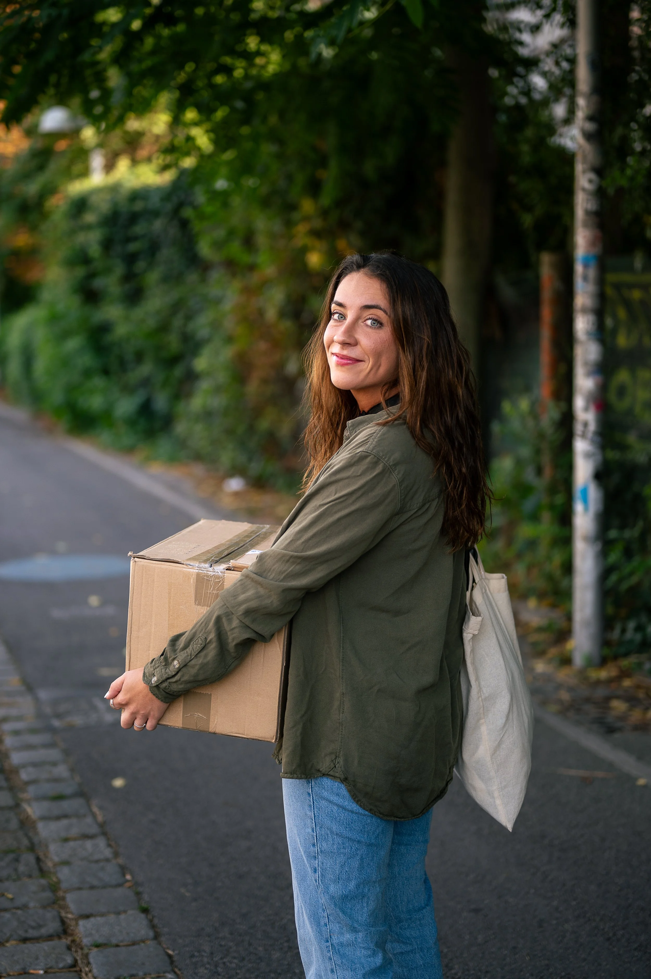 A woman with long brown hair standing on a sidewalk holding a cardboard box, wearing a green jacket and blue jeans, with a tote bag over her shoulder, looking back and smiling.