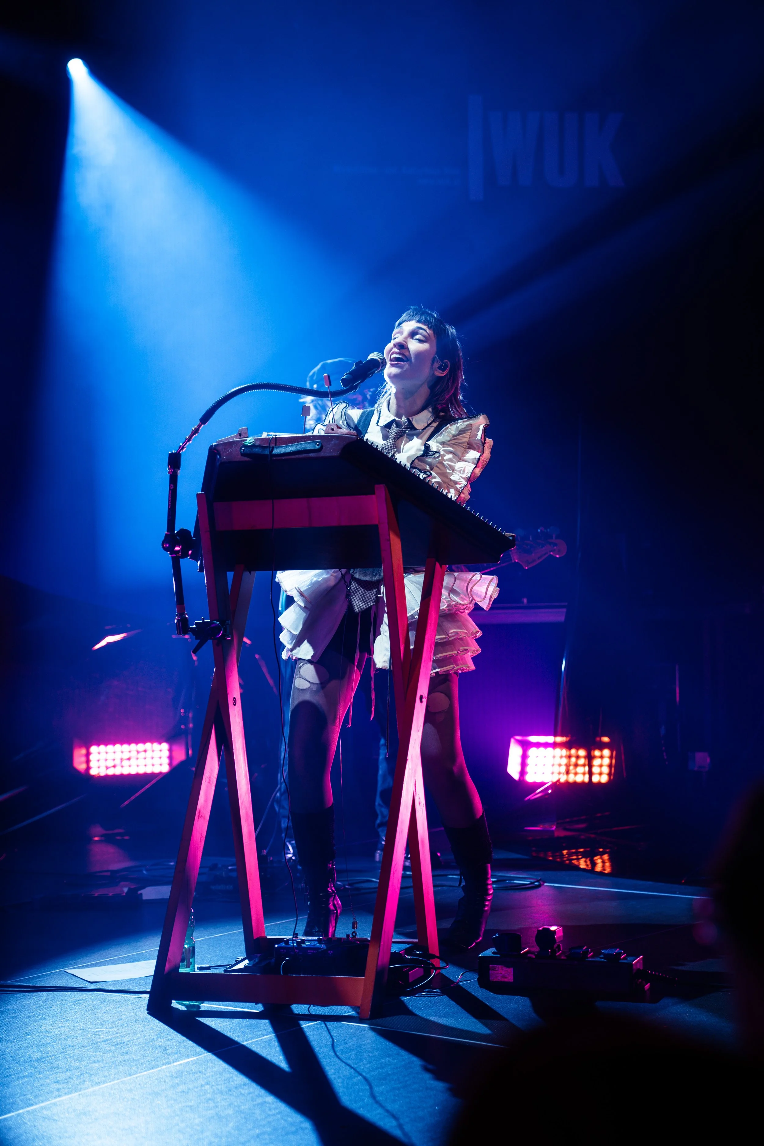 A woman performs on stage, singing into a microphone while playing an electronic keyboard, with stage lighting creating a vibrant atmosphere.