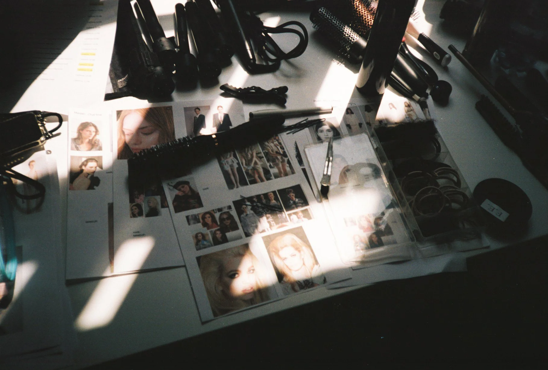 A photography or makeup station with various photos of models, makeup tools, and accessories scattered on a white table, partially illuminated by sunlight.