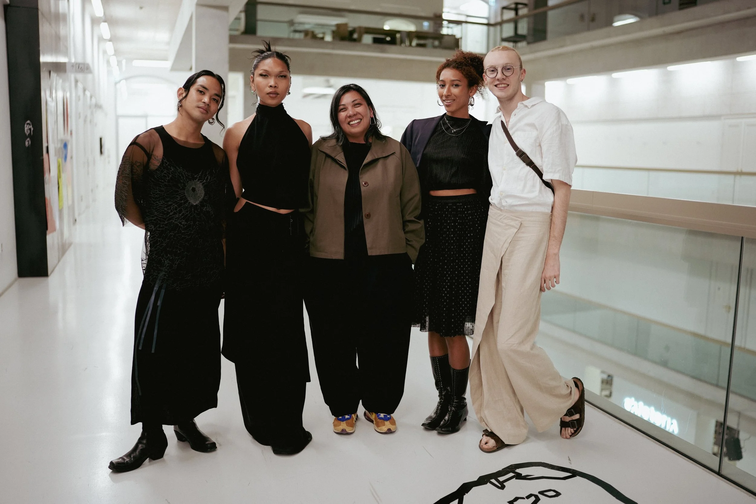 A diverse group of five women standing together on a balcony in a modern building, smiling at the camera.