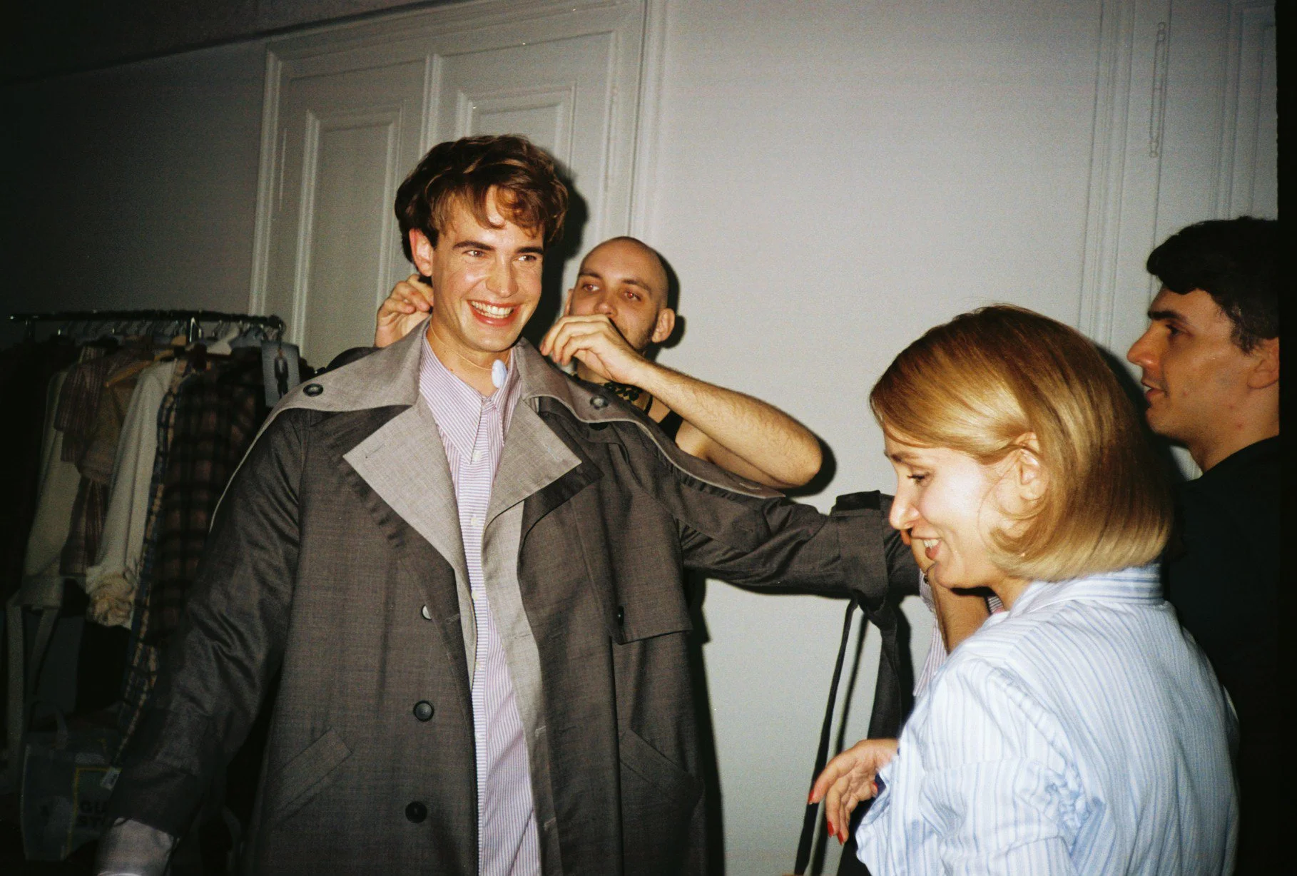 People in a dressing room, one man trying on a coat, others helping with the fitting, smiling and chatting.