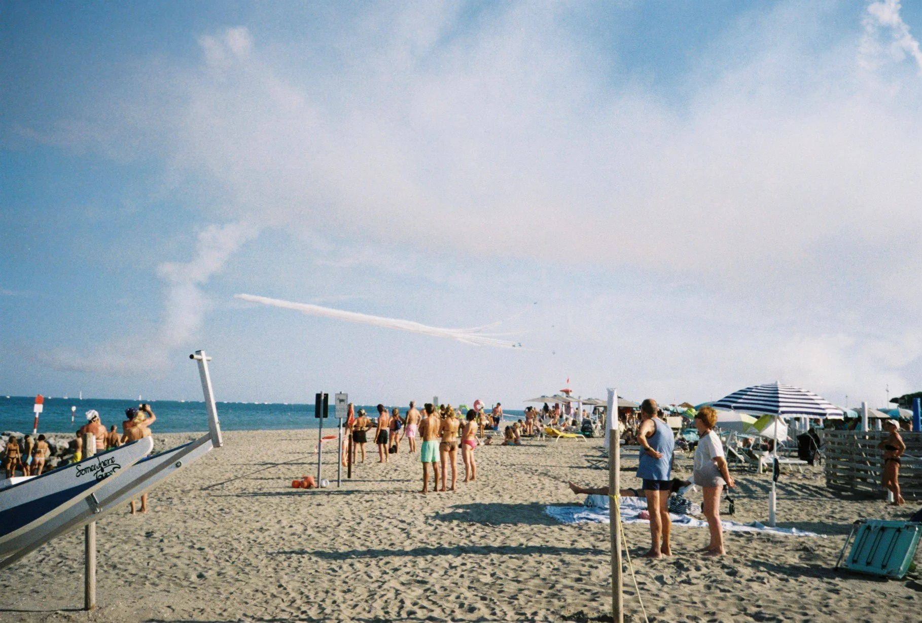 A busy beach scene with people sunbathing, swimming, and playing volleyball under a partly cloudy sky, with umbrellas and beach chairs along the sandy shore.