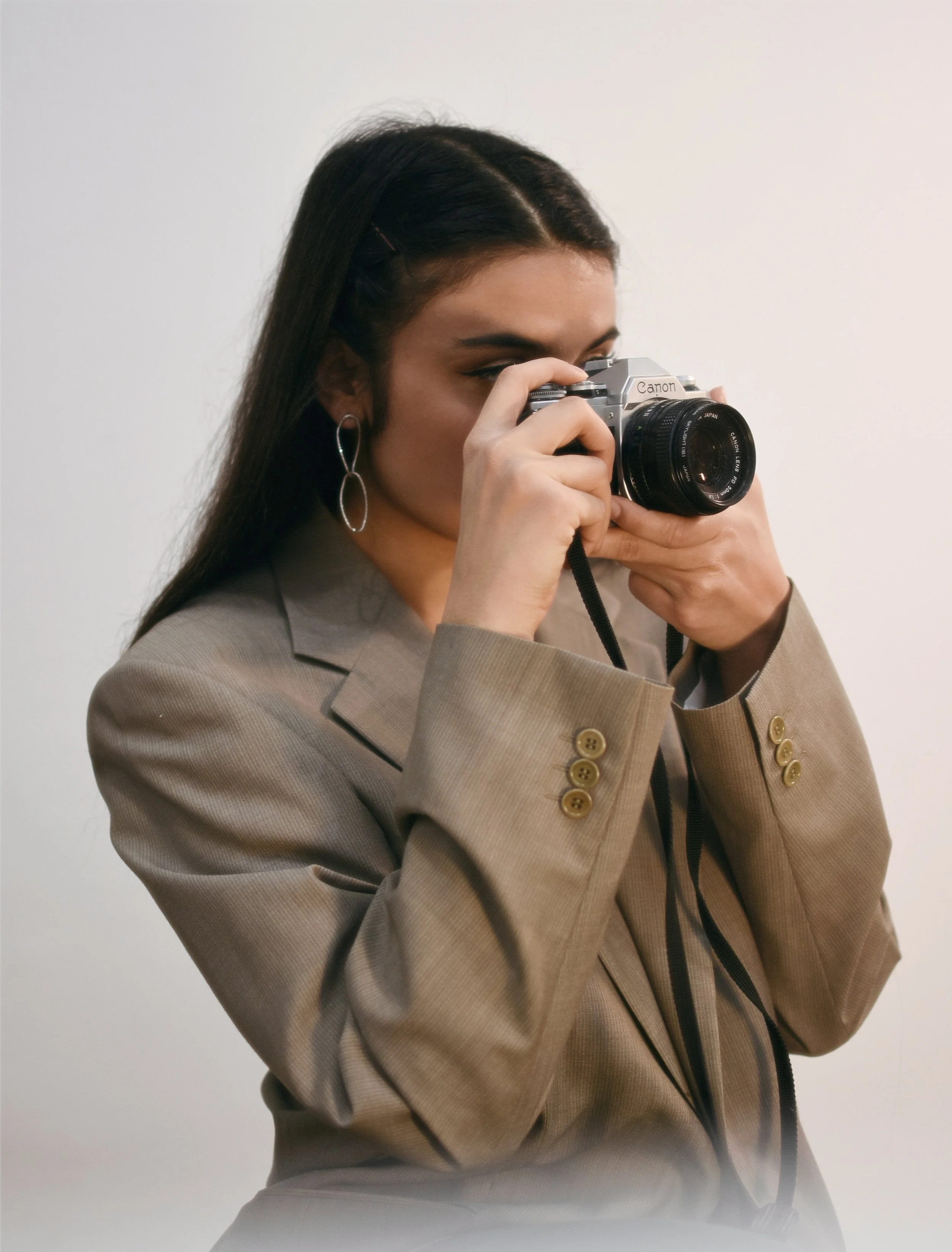 A woman with dark hair and large earrings wearing a tan blazer, holding a camera up to her face, about to take a photograph.