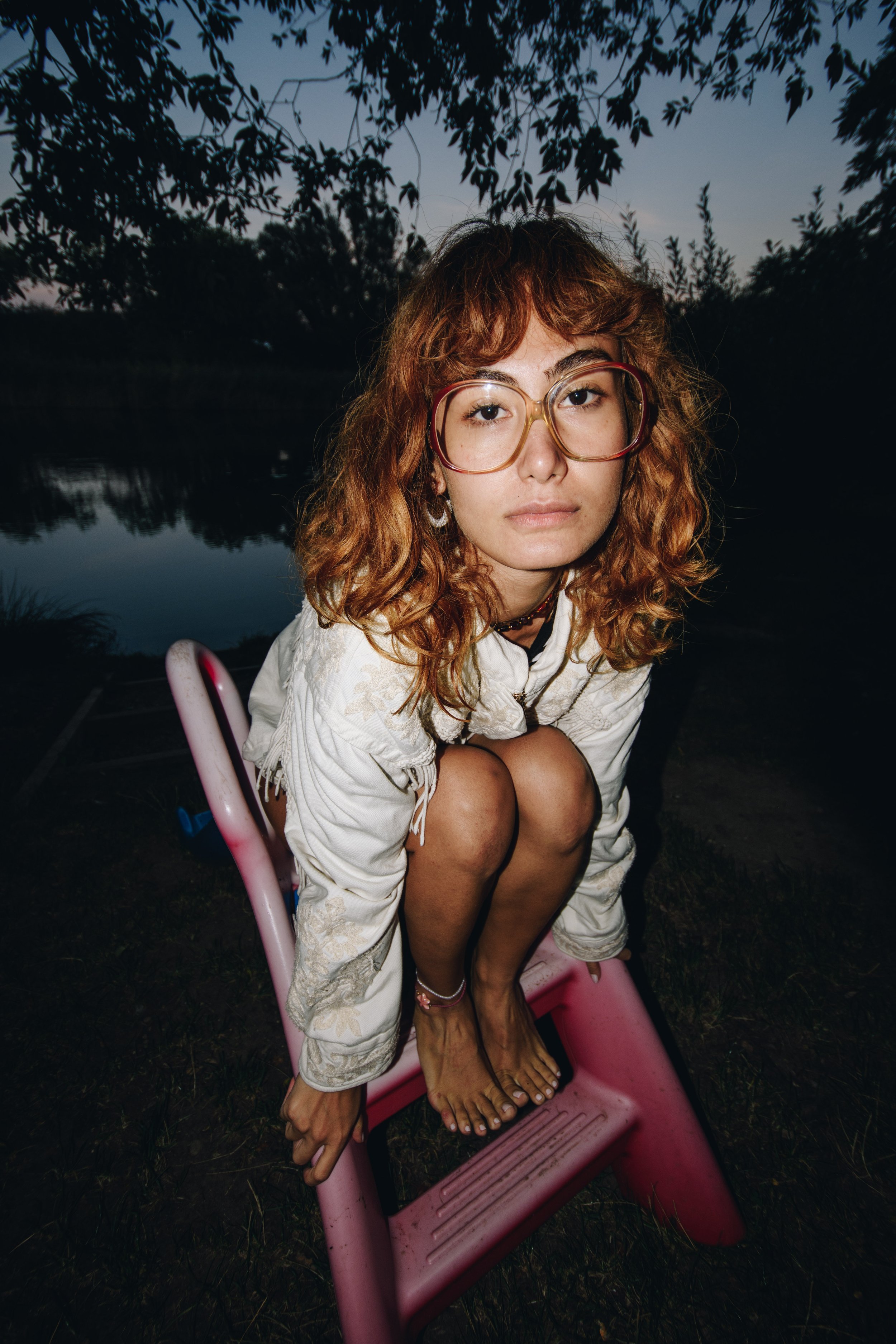 A young woman with red curly hair and large glasses crouches on a pink plastic chair outdoors at dusk near a body of water, with trees and a dark sky in the background.