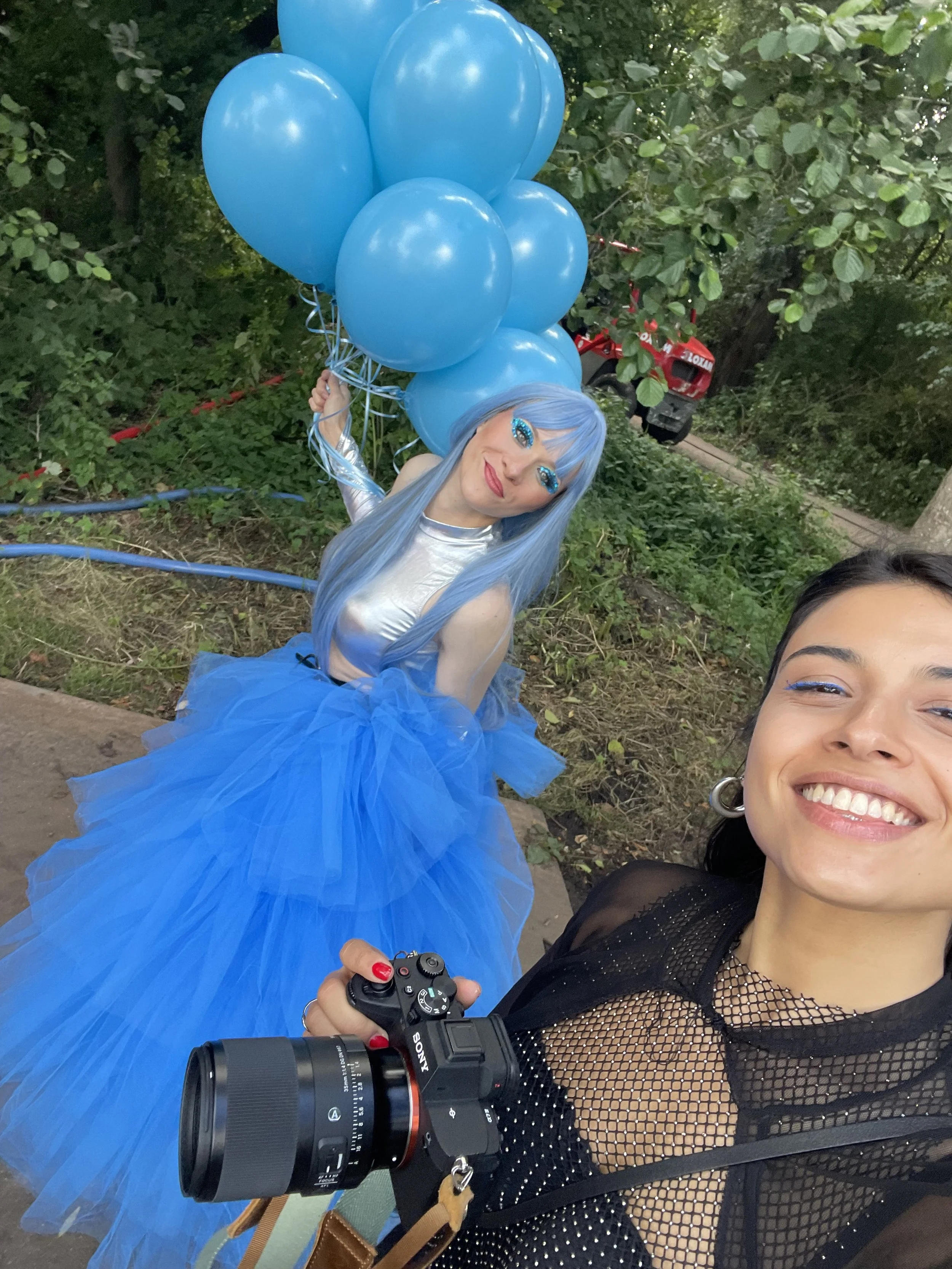 A woman with black hair and hoop earrings taking a selfie with a woman dressed in a blue outfit and holding blue balloons in a green outdoor setting.