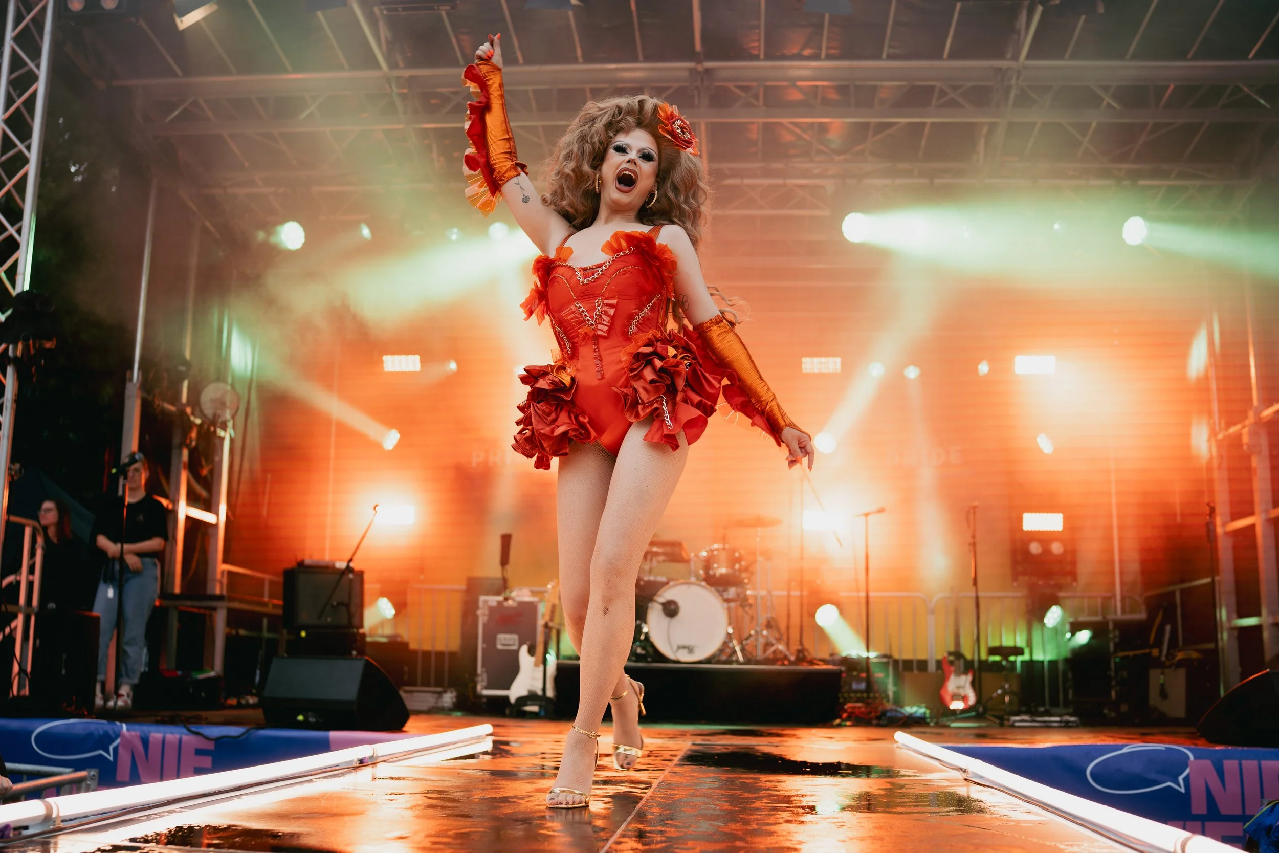 Performer in a vibrant red costume singing on stage with bright stage lighting and musical instruments in the background.