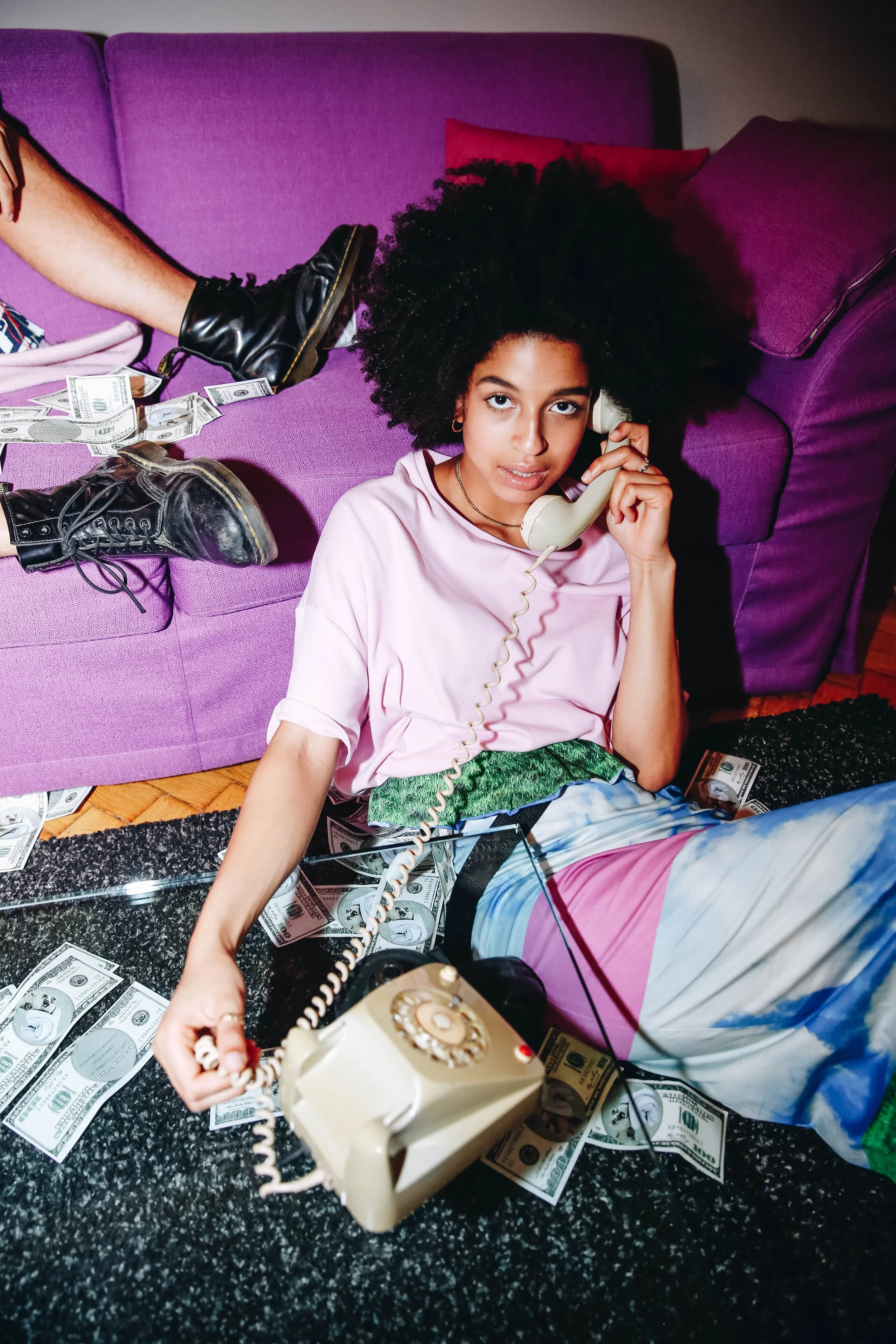 Young woman with a large curly hairstyle, sitting on the floor with money scattered around her, talking on a corded landline phone, in a room with a purple sofa and colorful pillows.