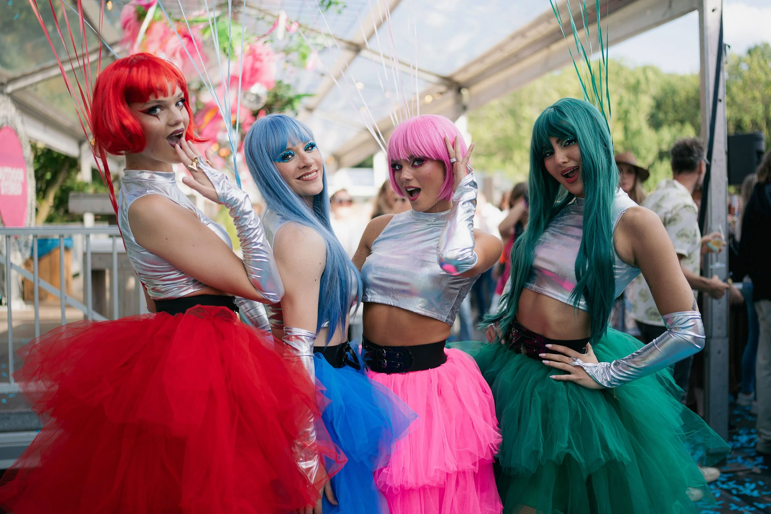 Four women dressed in colorful, shiny costumes with bright skirts and wigs, posing together at an outdoor event.
