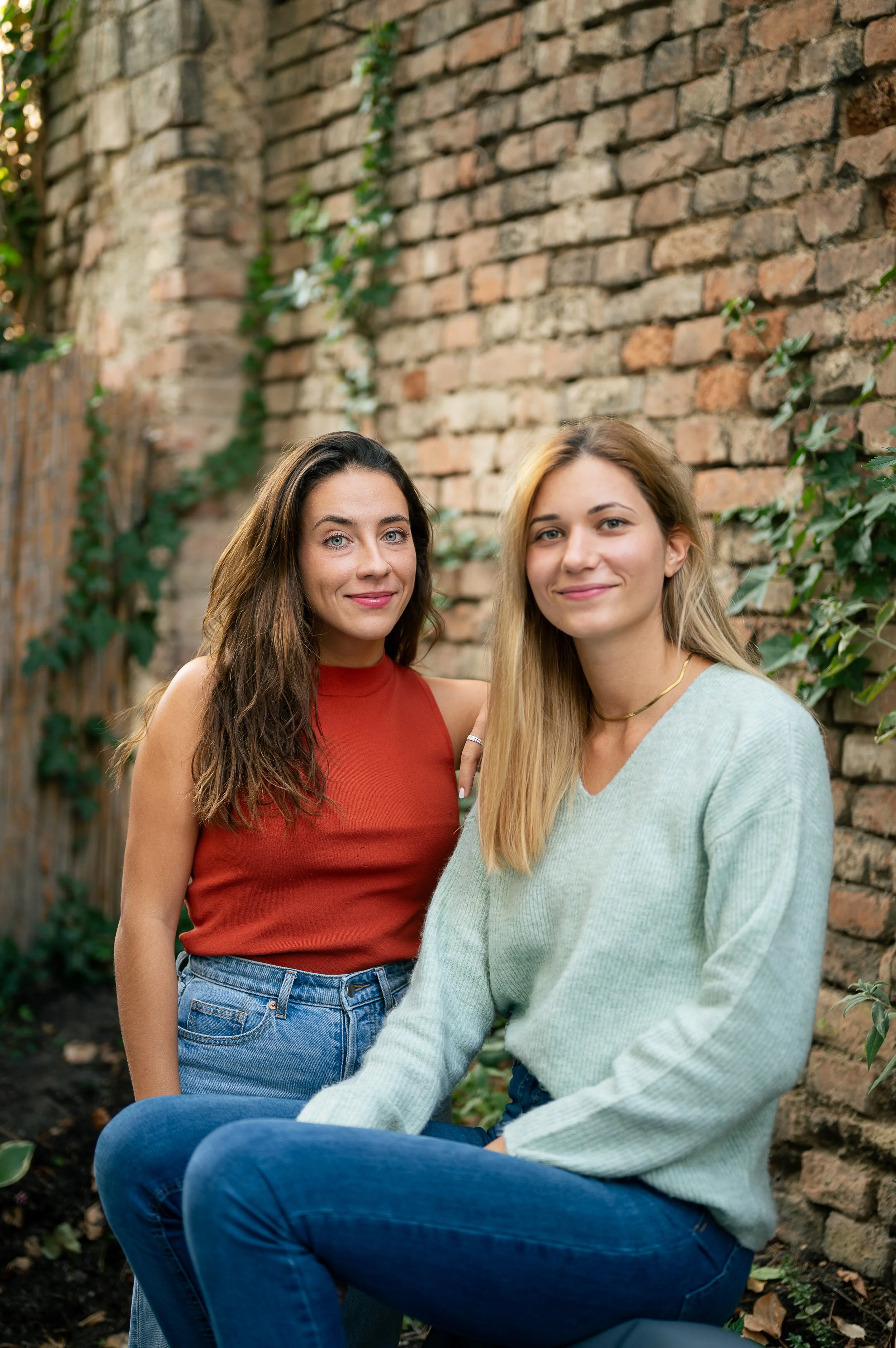 Two women sitting outdoors near a brick wall with green ivy. One woman has long dark hair, wearing a sleeveless rust-colored top and blue jeans. The other woman has long blonde hair, wearing a light green sweater and blue jeans.