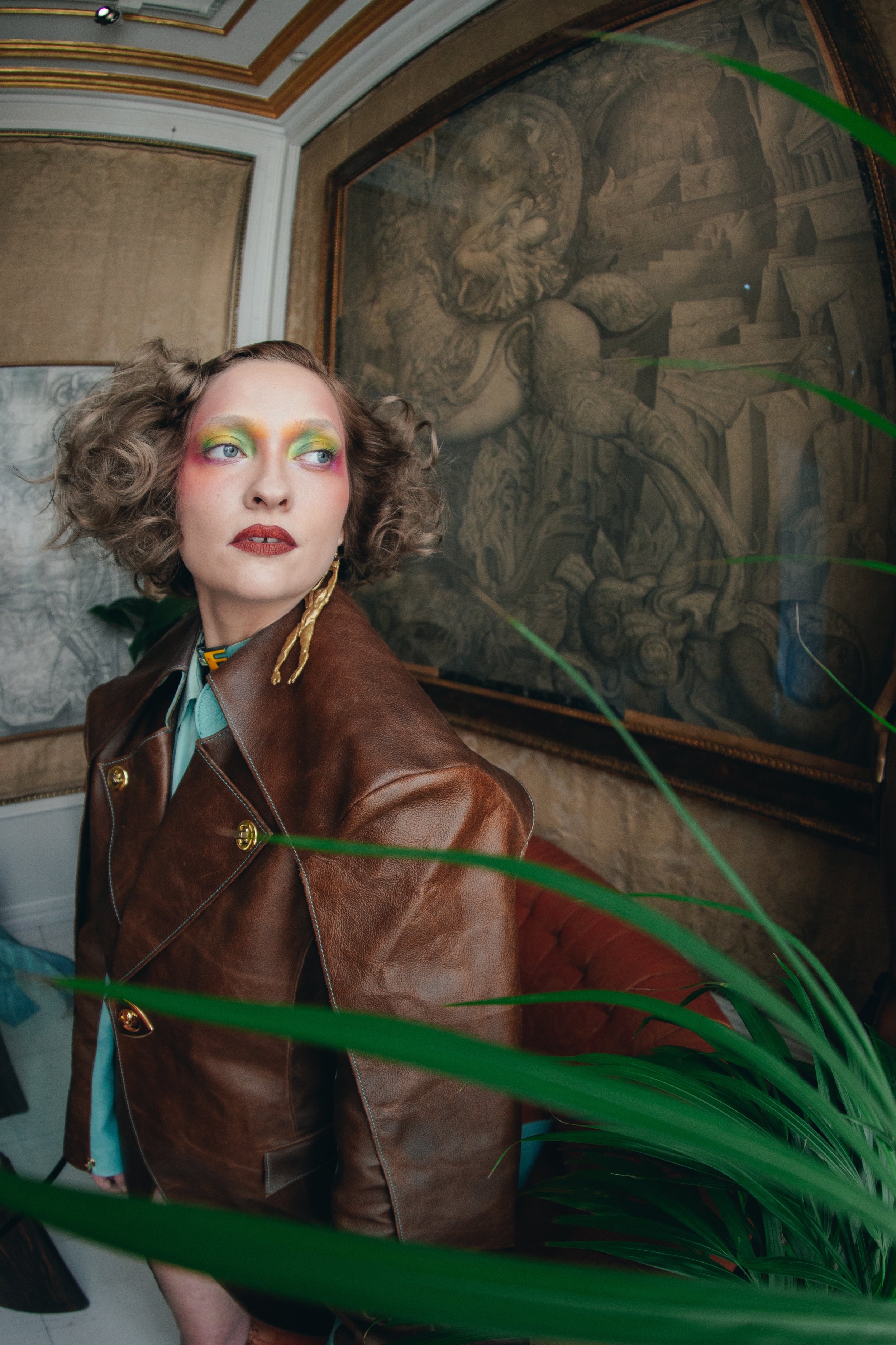 A woman with colorful eye makeup and curly hair in an art gallery, wearing a brown leather jacket, seated near paintings and greenery.