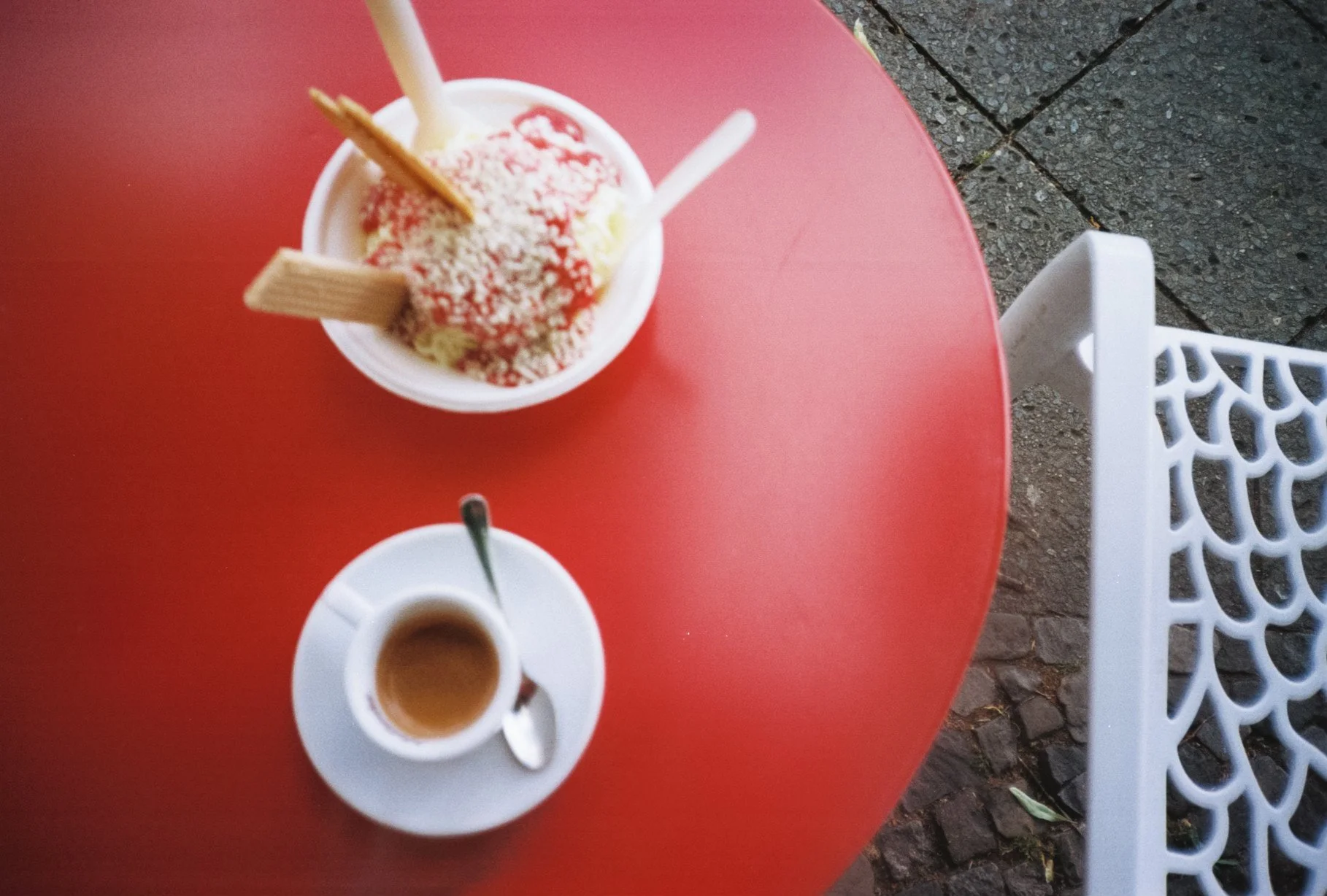 Ice cream sundae topped with shredded coconut and cookies on a red table, with a cup of coffee on a white saucer with a spoon.