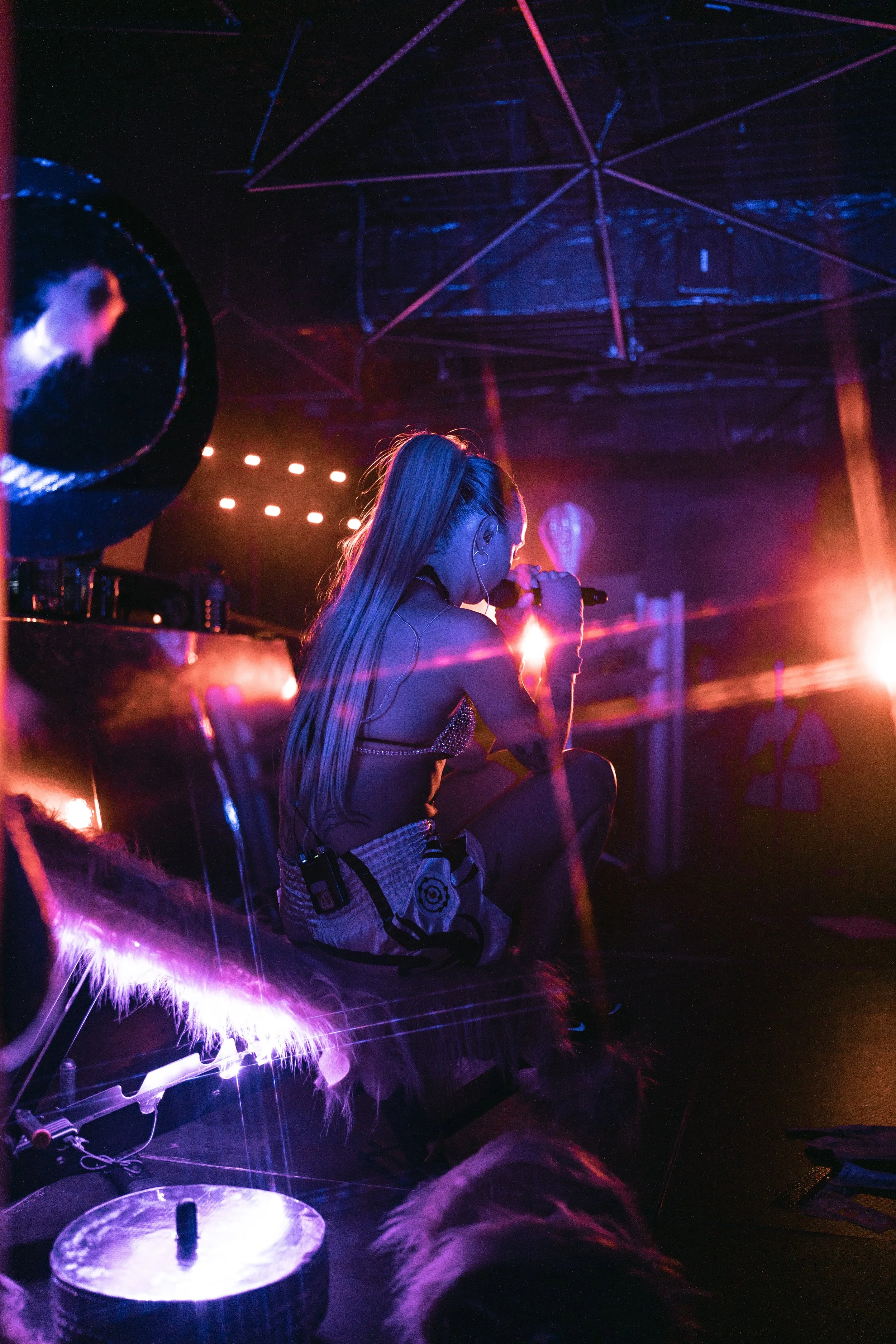 A performer with long blonde hair singing into a microphone on stage illuminated by colorful purple, pink, and orange lights in a dark nightclub or concert venue.