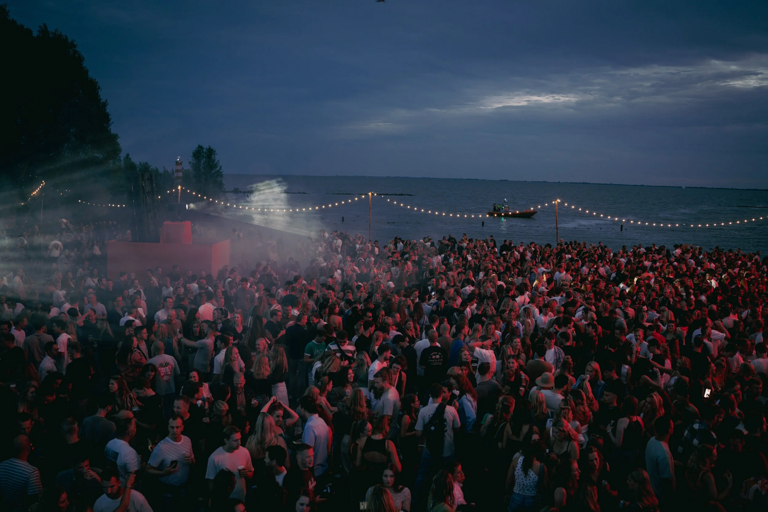 Large crowd of people gathered on a beach at night, with string lights hanging and a boat on the water in the background.