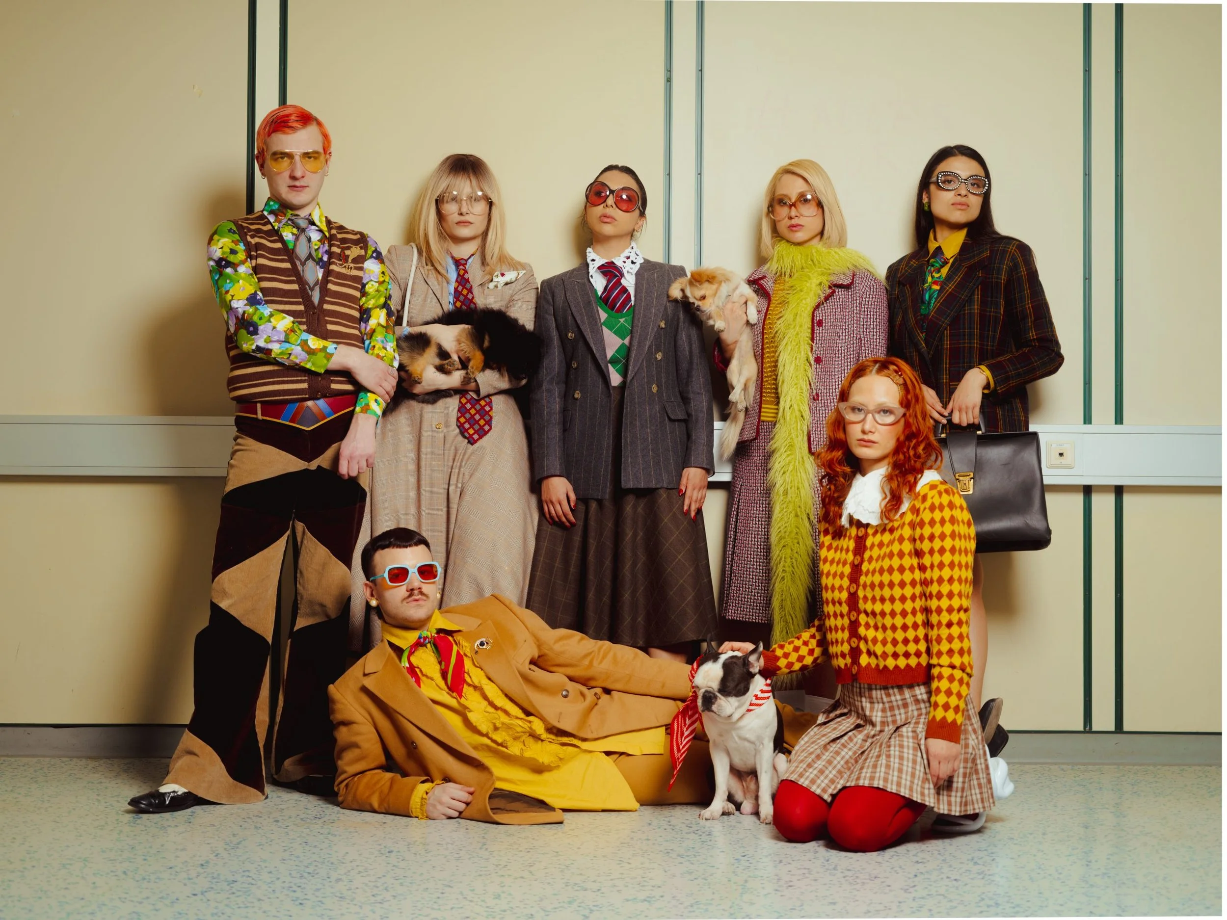 A group of nine people dressed in colorful, vintage-inspired outfits, some holding pets, standing and sitting in front of a beige wall.