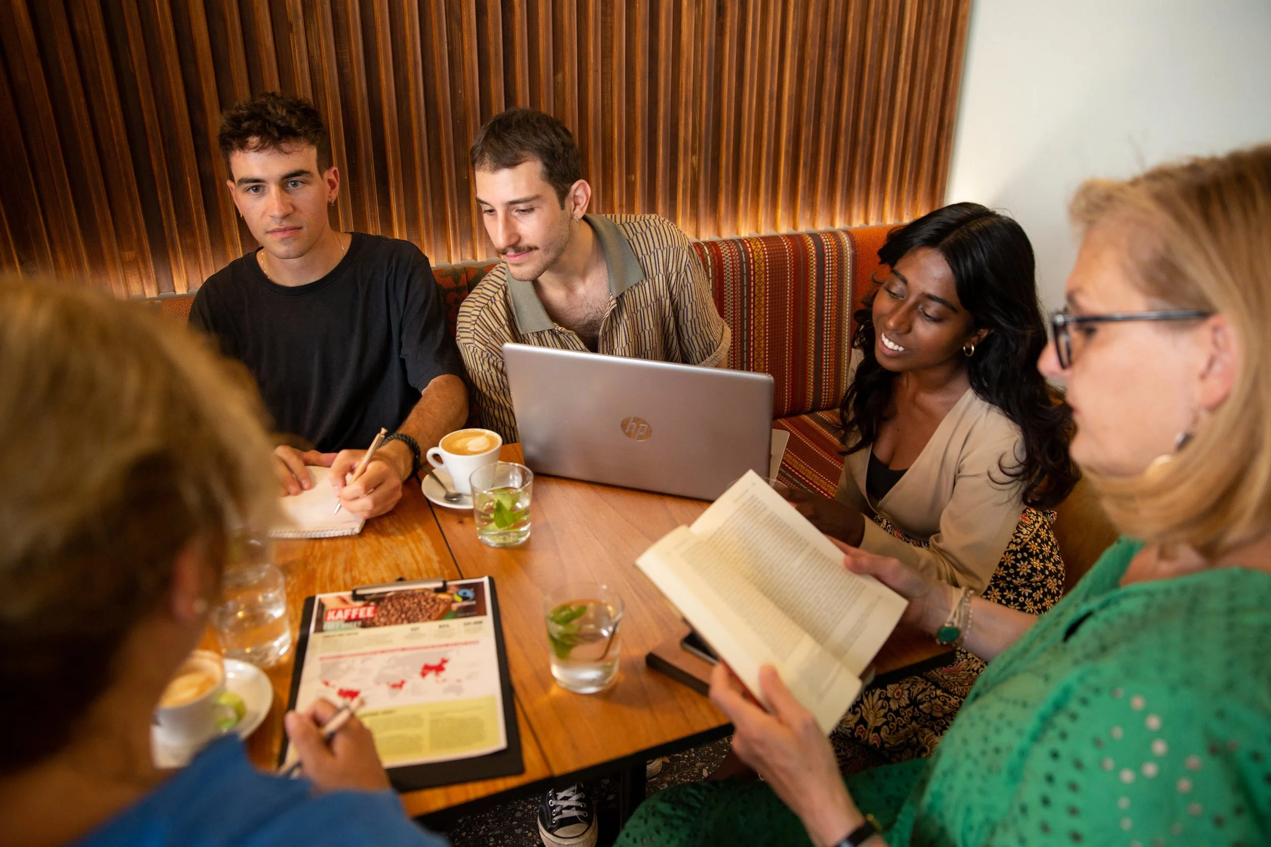 Group of six people sitting around a wooden table in a cafe, engaged in discussion. There are coffee cups, glasses of water, a laptop, and printed materials on the table.